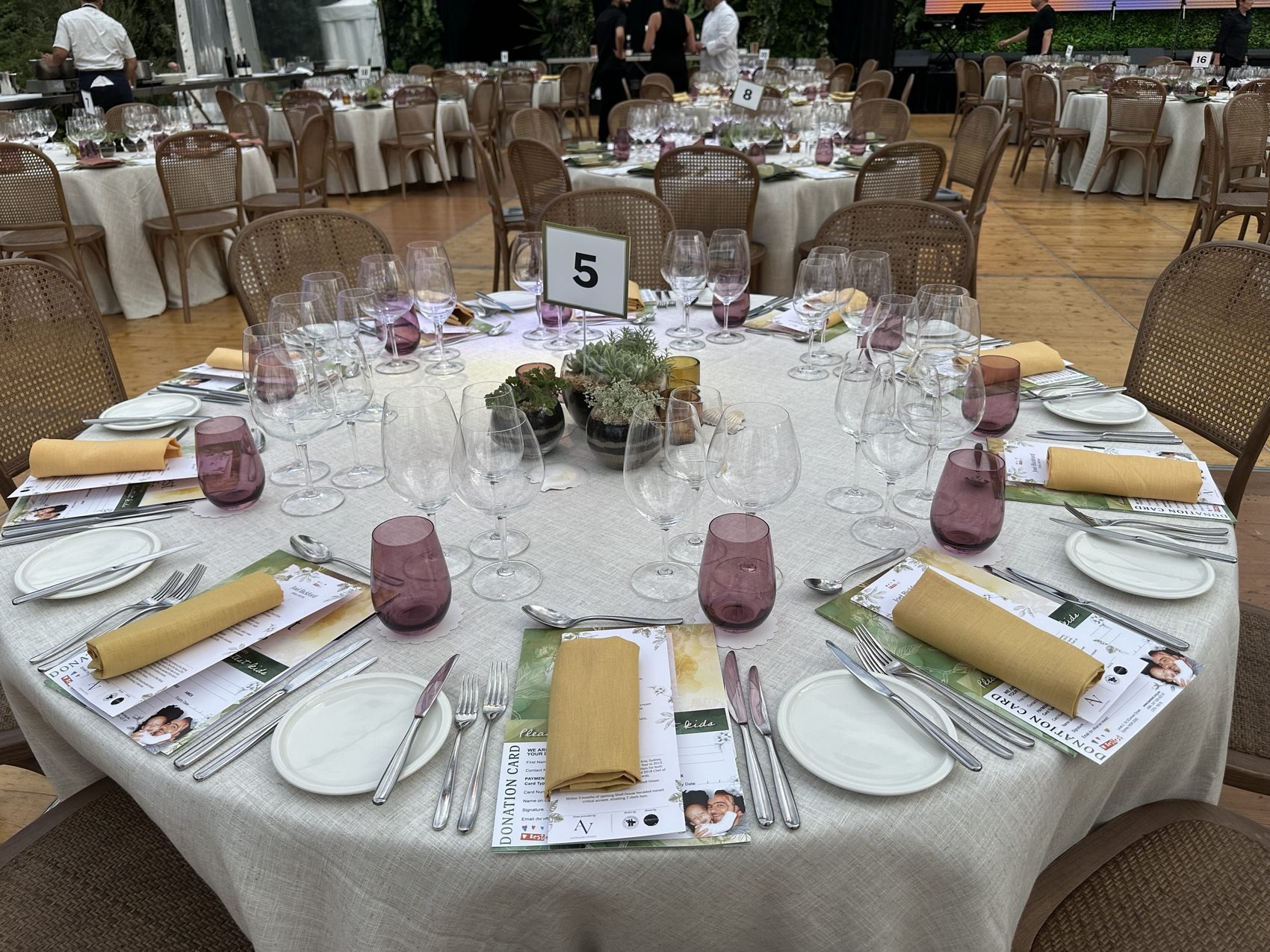 A round banquet table set for a formal event, with wine glasses, plates, silverware, yellow napkins, and printed programs or menus. The table has a centerpiece with potted plants and is labeled with a table number sign '5'. Multiple similar tables are visible in the background, all arranged in a decorated event space.