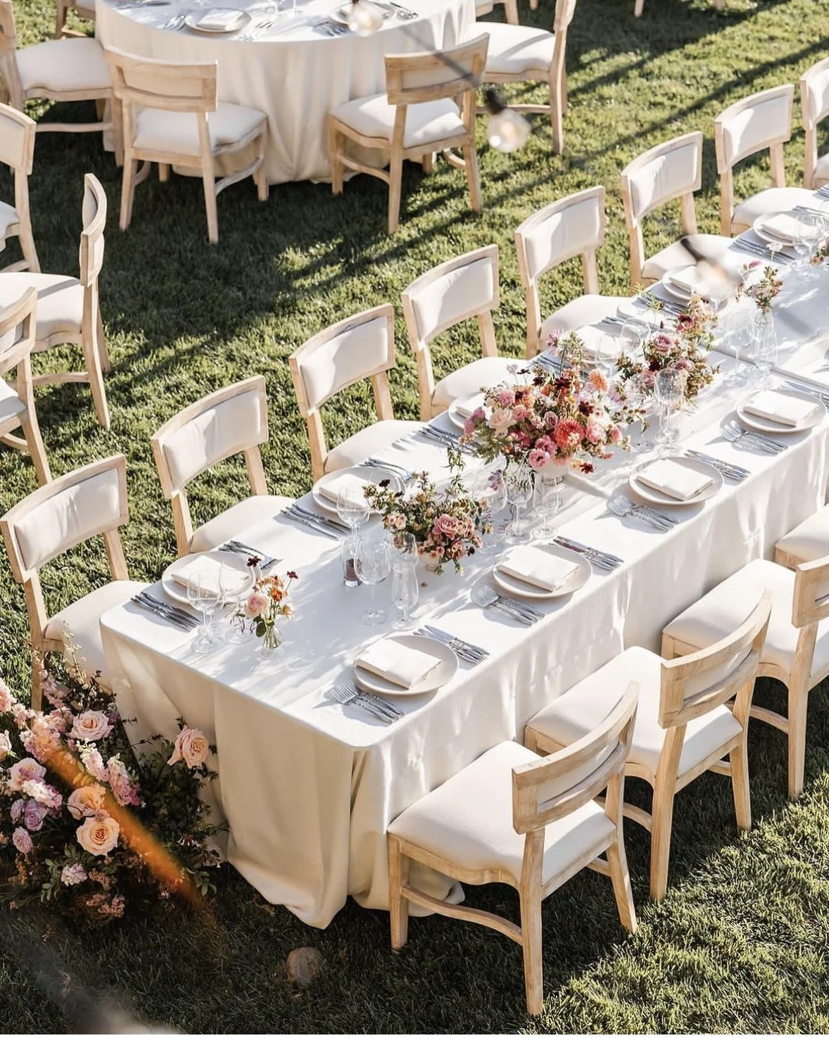 Elegant outdoor event table setup on grass with white tablecloth, floral centerpiece, and wooden chairs.