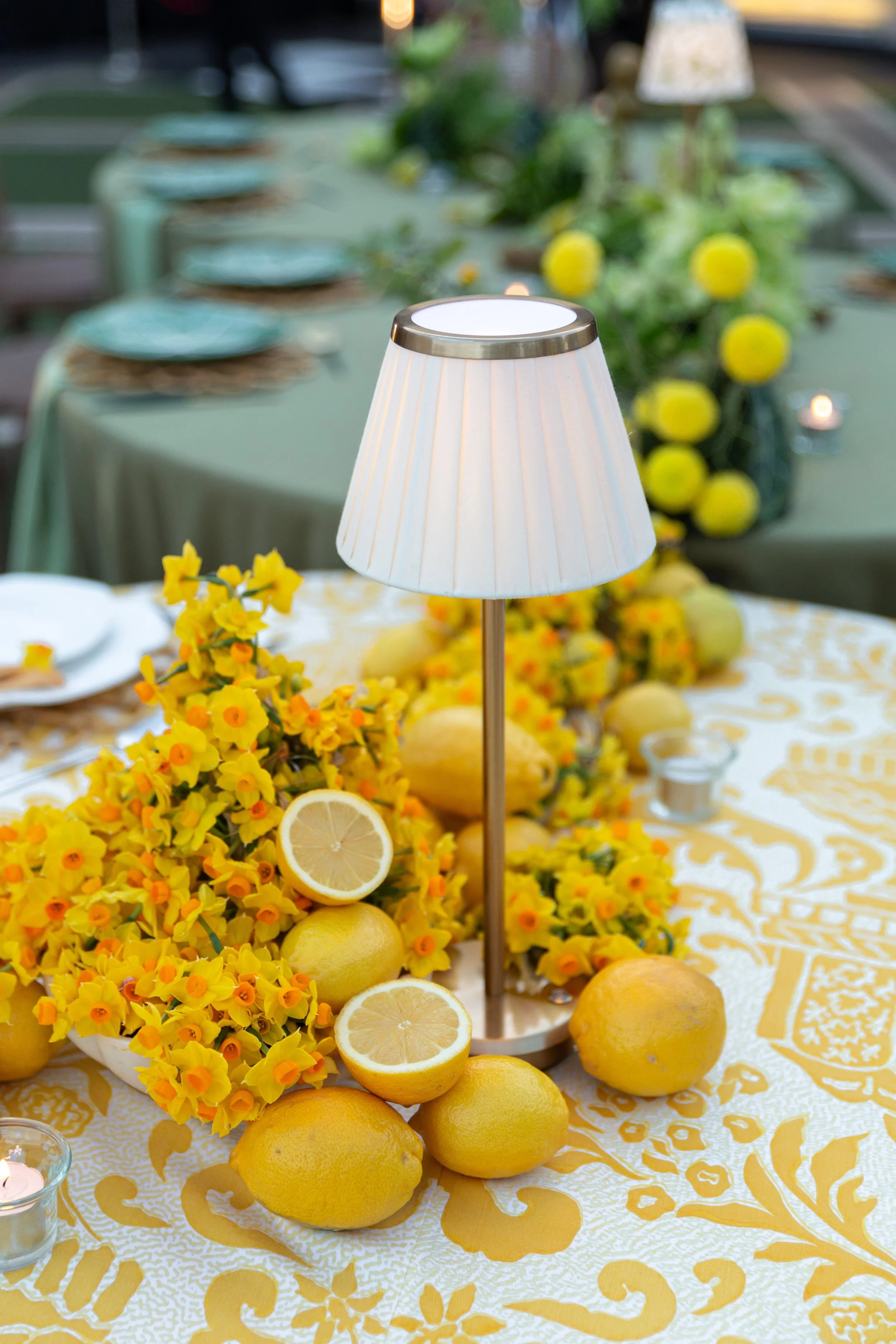 A table decorated for a celebration with yellow lemons, yellow flowers, and a centerpiece featuring a table lamp. The tablecloth and plates are in coordinated colors.