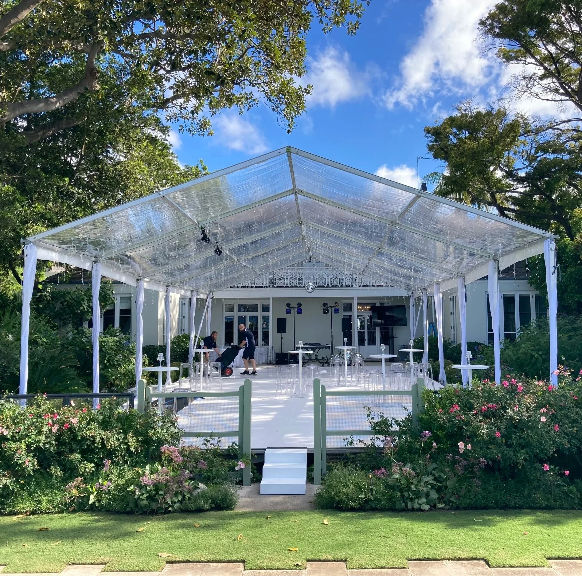 Outdoor event space setup with a white canopy tent decorated with hanging fairy lights, surrounded by lush greenery and blooming flowers, with some people setting up inside under a clear blue sky.