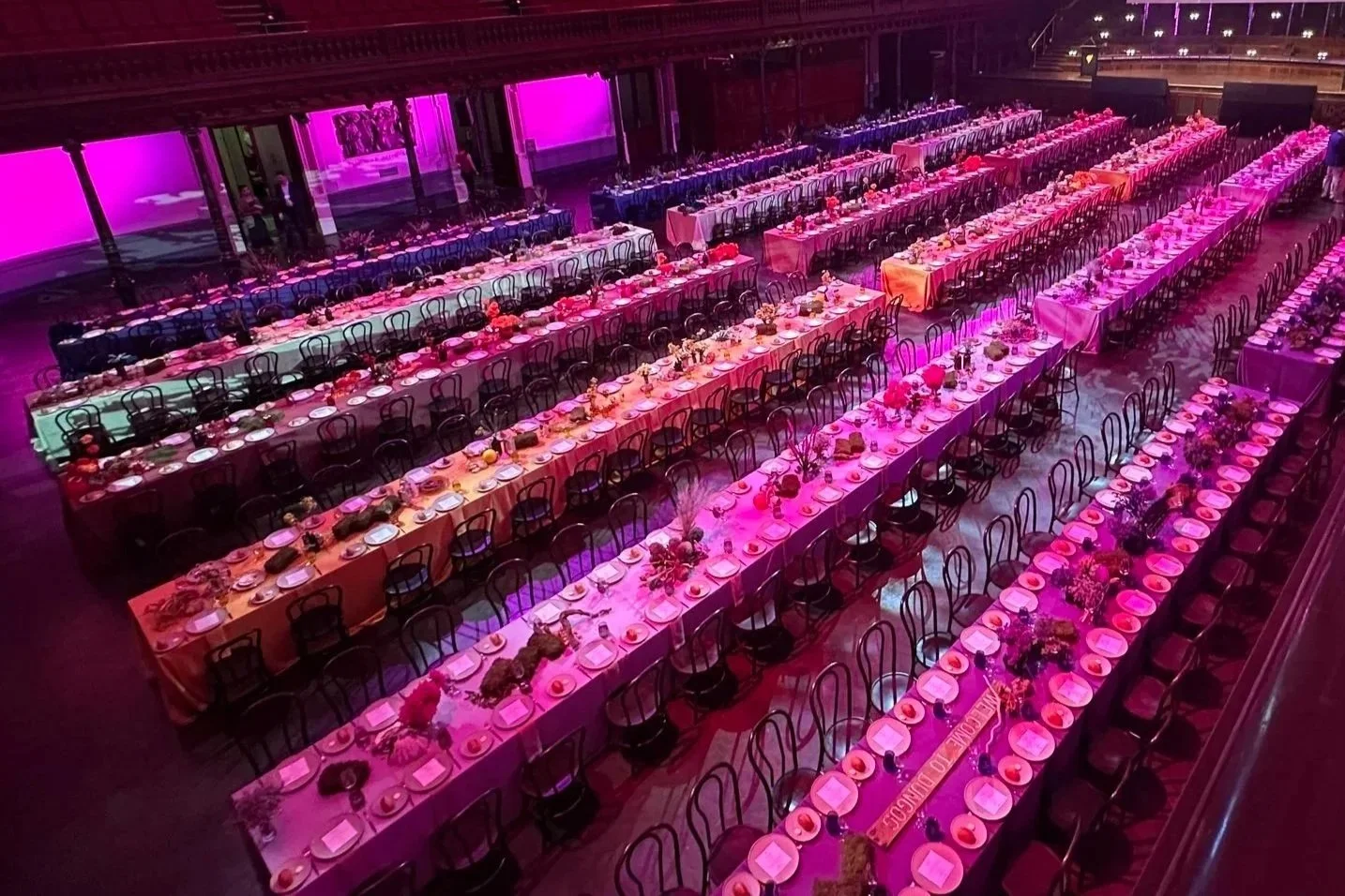 Large banquet hall decorated with long tables set with plates, glasses, and centerpieces, illuminated by pink and purple lighting.