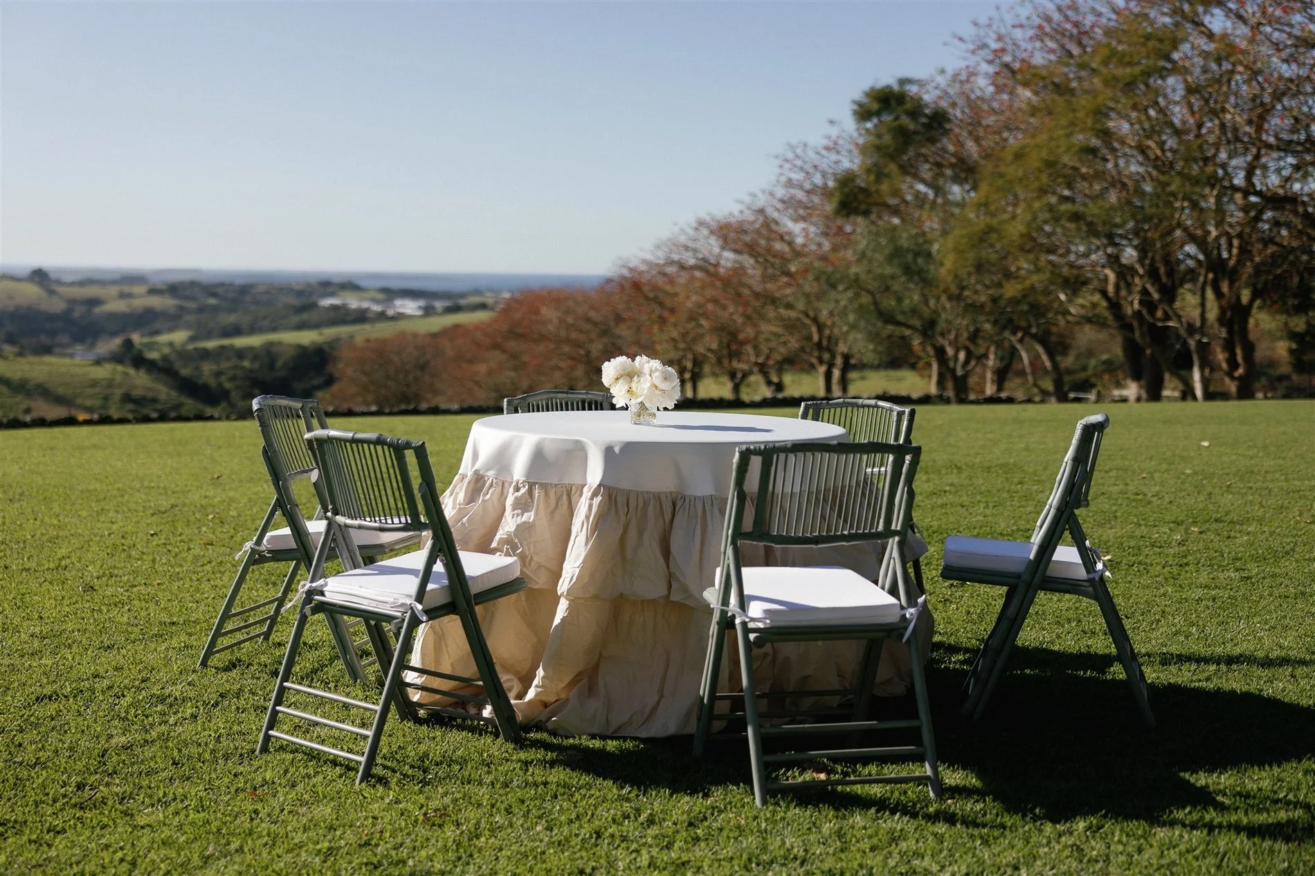 Outdoor round table with a white tablecloth and a floral centerpiece, surrounded by six gray folding chairs on a grassy field with trees in the background.