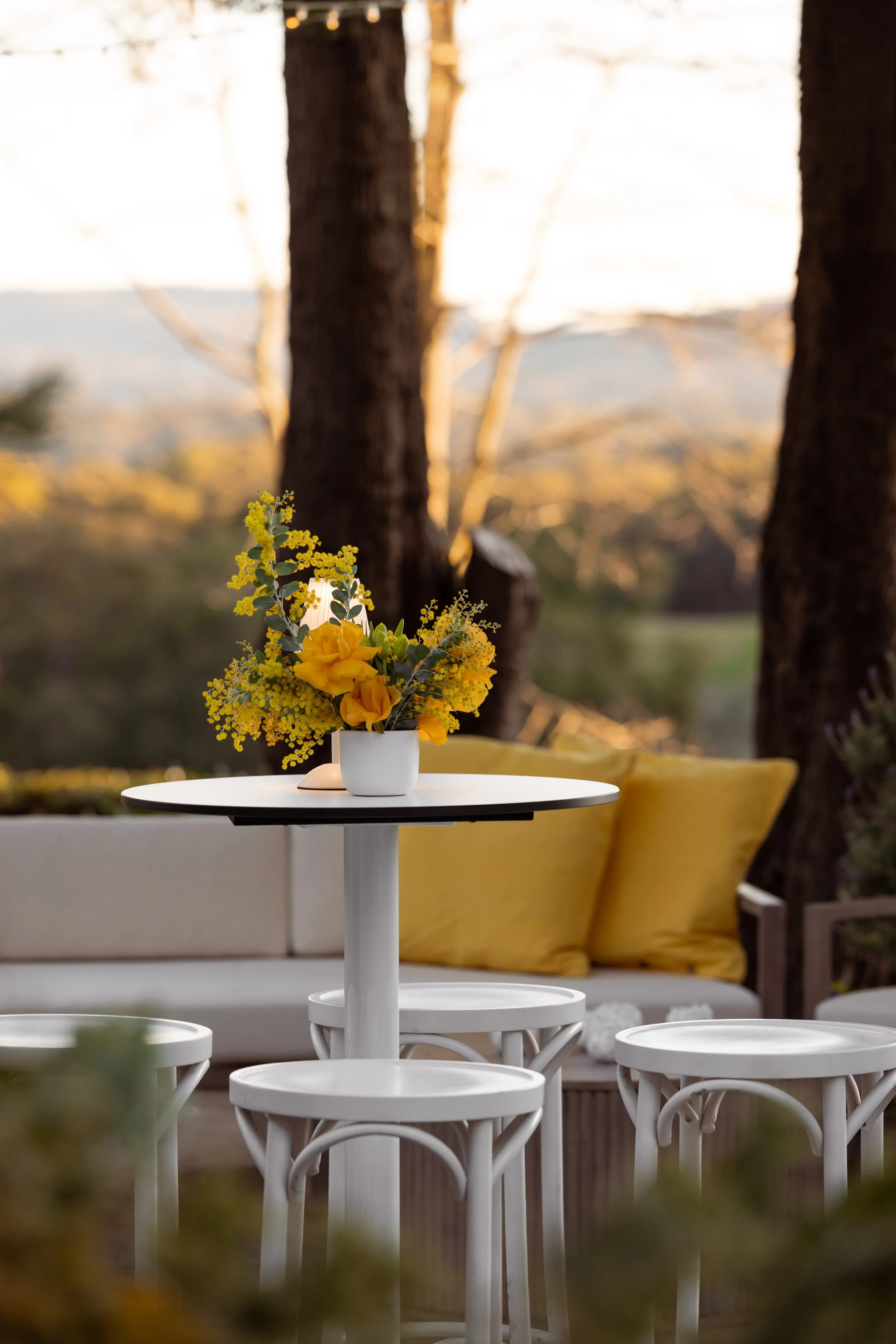 A white round table with a white flower vase containing yellow flowers. A white cushioned bench with yellow pillows in the background. Trees and a sunset in the distance.