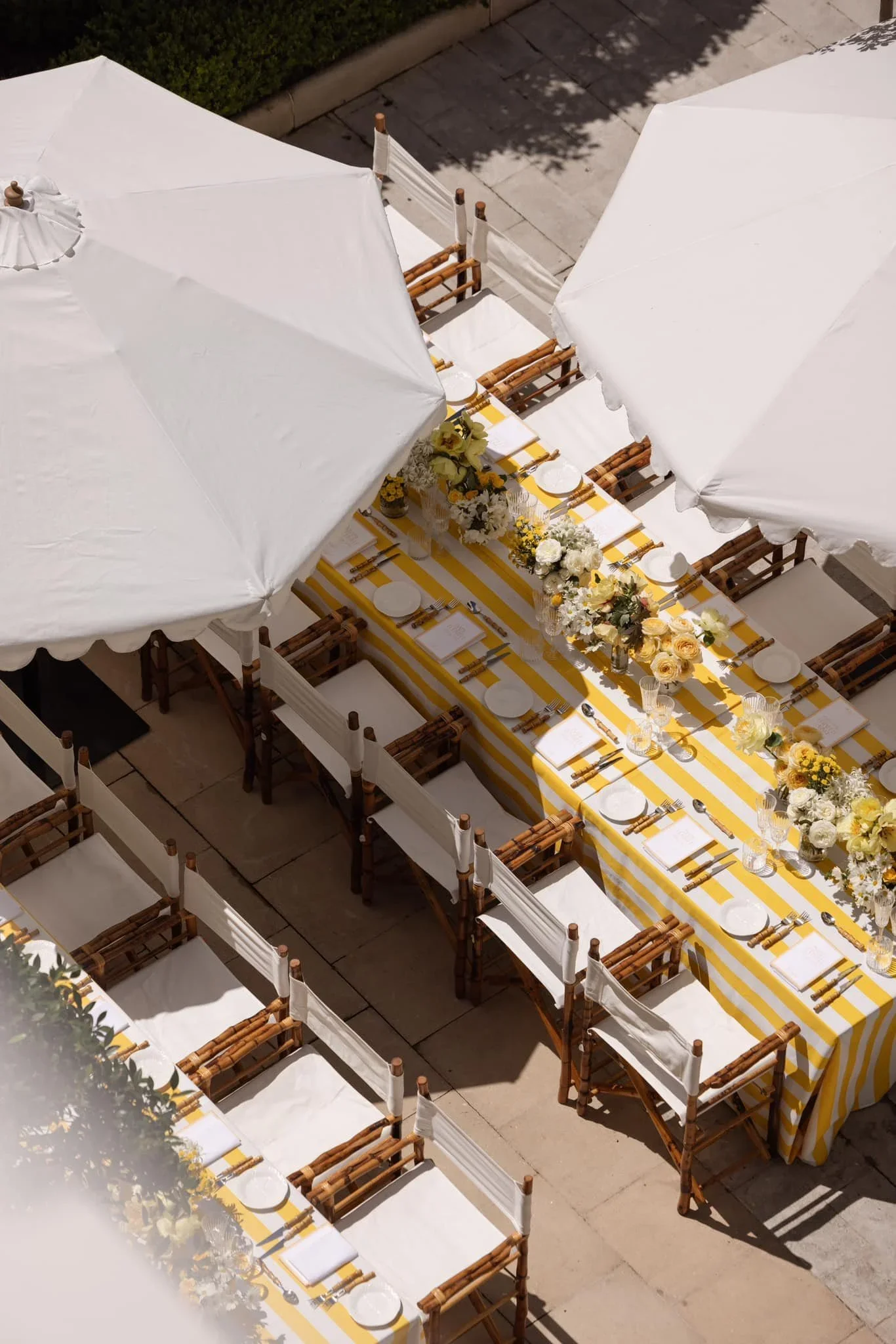 Outdoor dining table set under white umbrellas with yellow and white striped tablecloth, floral centerpieces, plates, glasses, and silverware, on a paved patio.