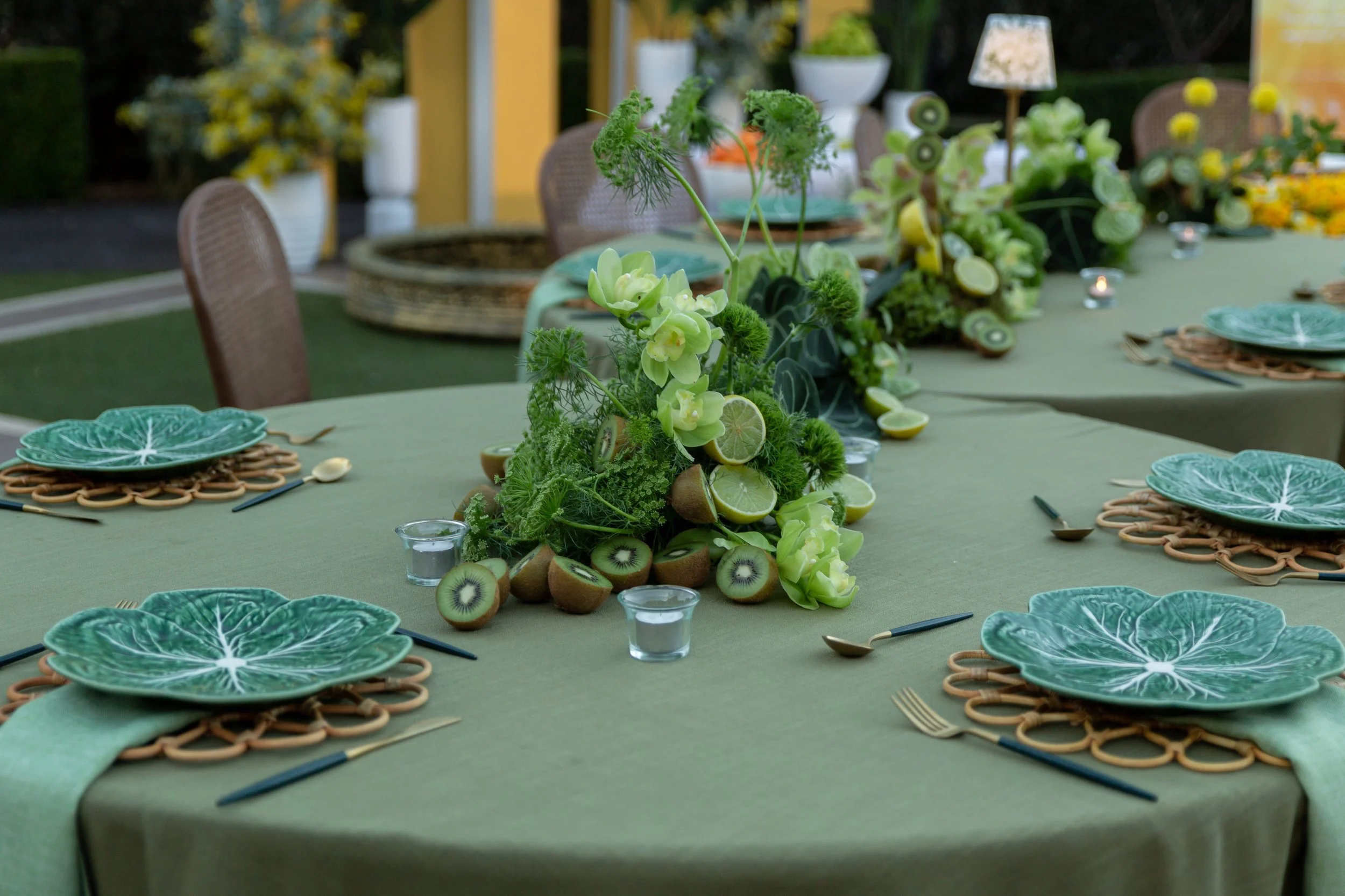 A dining table outdoors with a green tablecloth, set with teal leaf-shaped plates, gold utensils, small glass candle holders, and a floral centerpiece made of green flowers, kiwi slices, limes, and greenery.