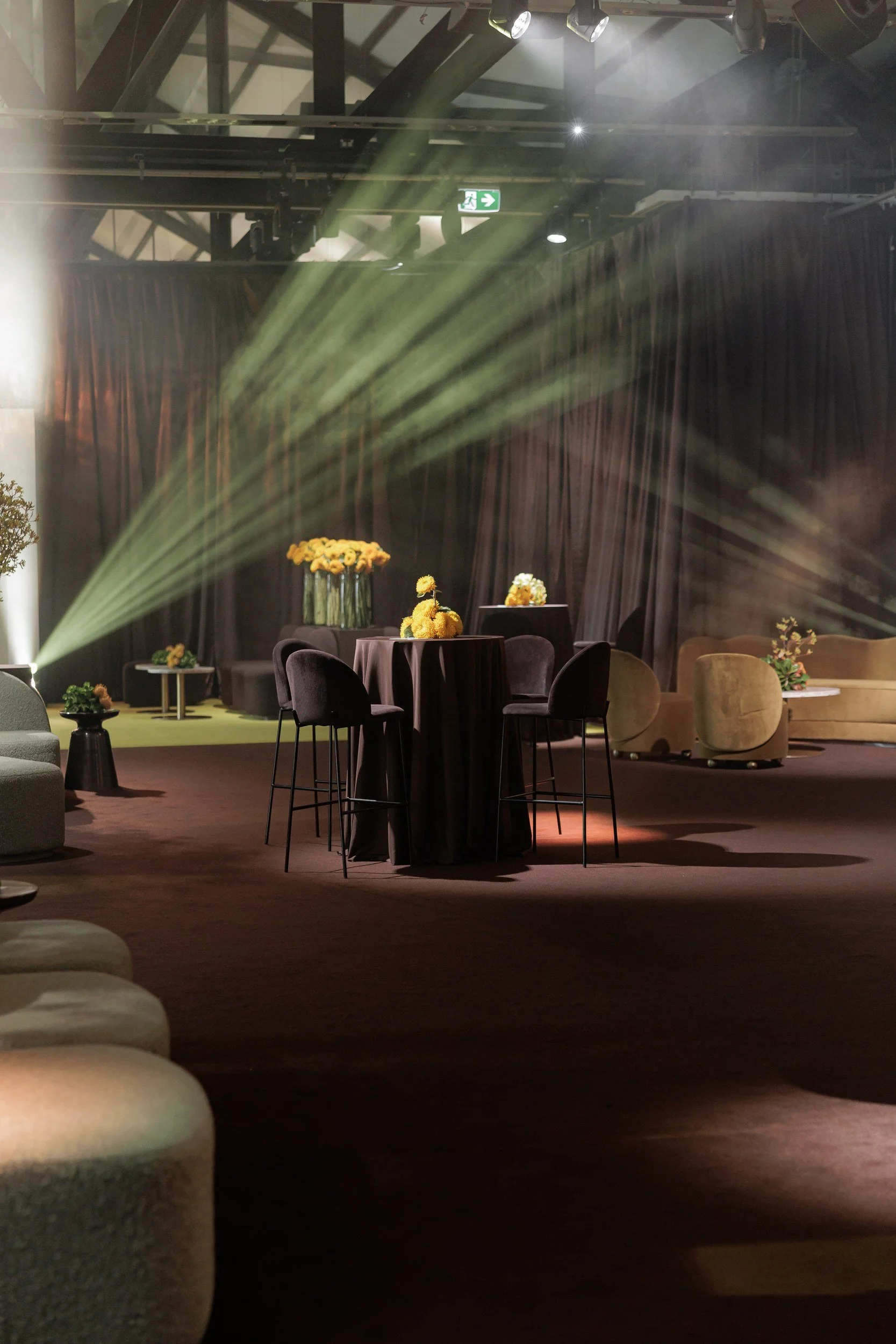 An indoor event space with a stage and seating area. Round tables decorated with yellow flowers and vases. Stage with curtains, greenery, and flower arrangements. Overhead lighting, a visible exit sign, and some dust or fog effects.