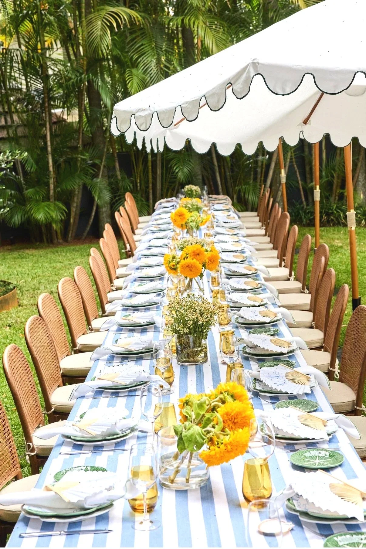 Long outdoor dining table set with floral centerpieces, place settings, and glasses under a white umbrella, surrounded by greenery.
