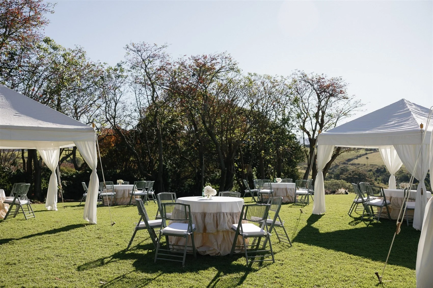 Outdoor event setup with round tables covered in white tablecloths and surrounded by chairs on a grassy lawn. White tents with draped curtains are set up, with trees and a hill in the background.