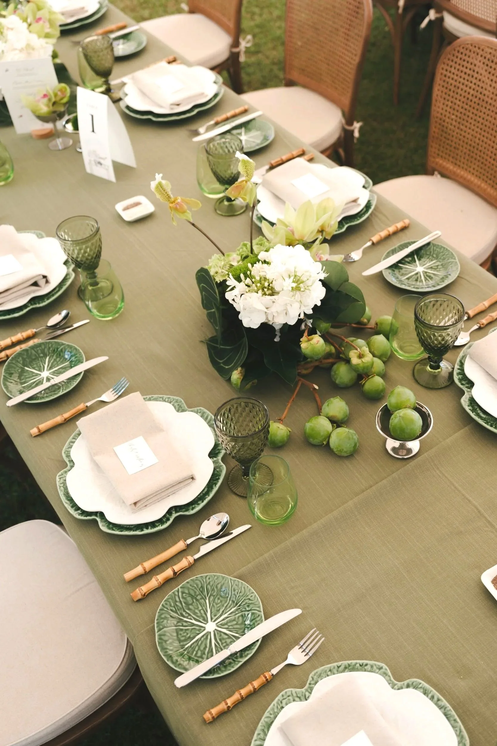 A decorated outdoor dining table set with green and white dishware, silverware, green glasses, napkins, and a floral centerpiece with white flowers and greenery.