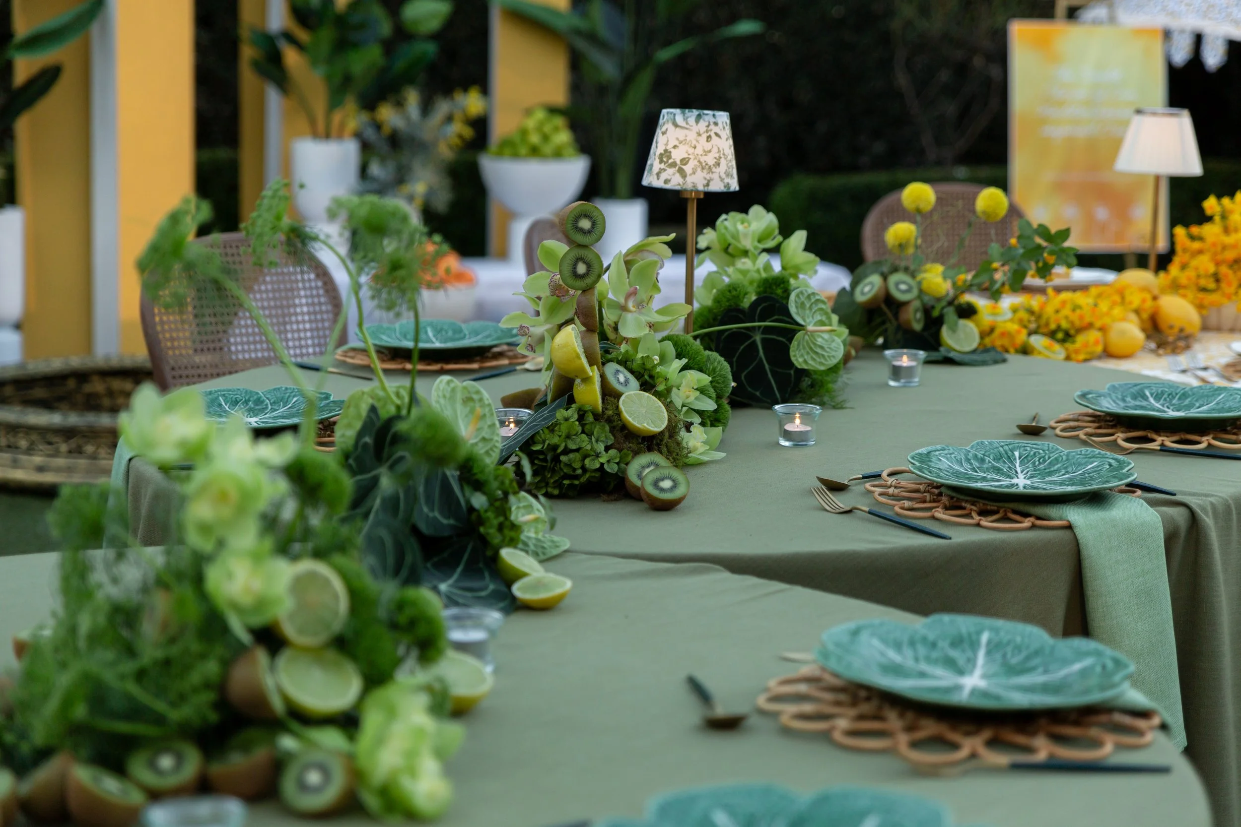 Decorated outdoor dining table with green tablecloth, floral centerpieces, paper lanterns, candles, and patterned plates, set in a garden with potted plants and greenery in the background.