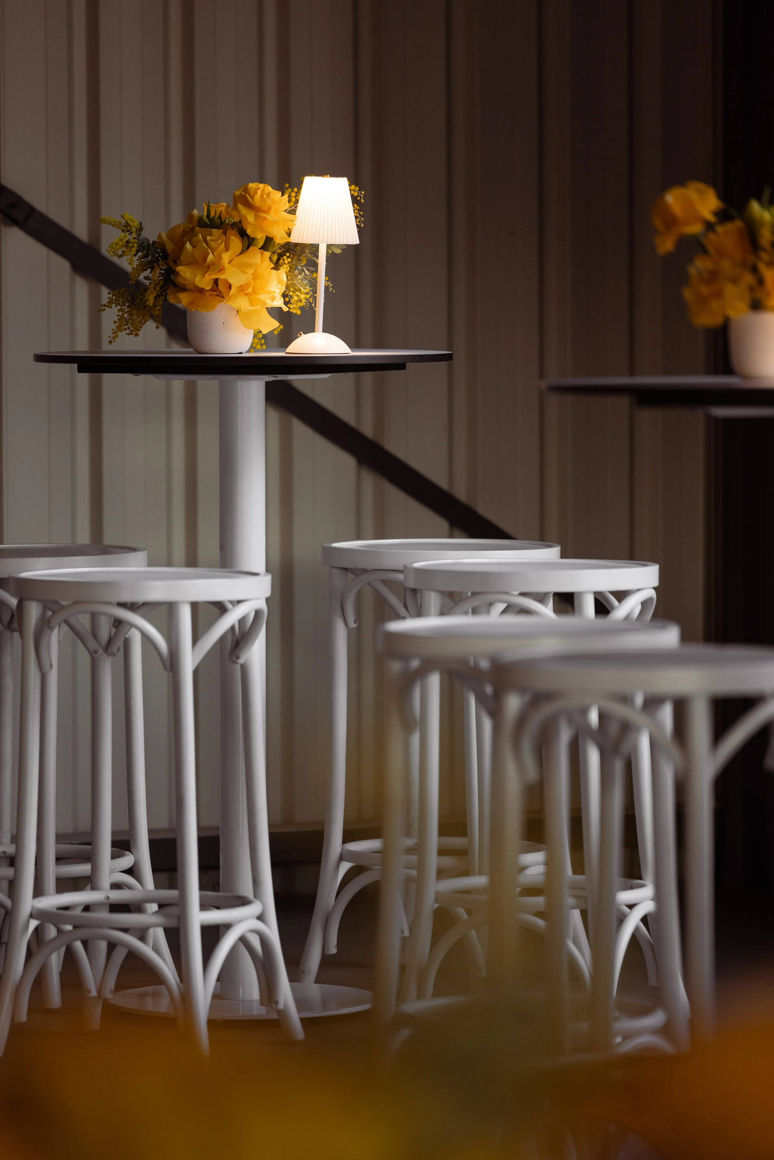Interior scene with white chairs arranged around a wooden table. On the table, there is a small lamp with a lampshade and a vase of yellow flowers. The background features wooden paneled walls.