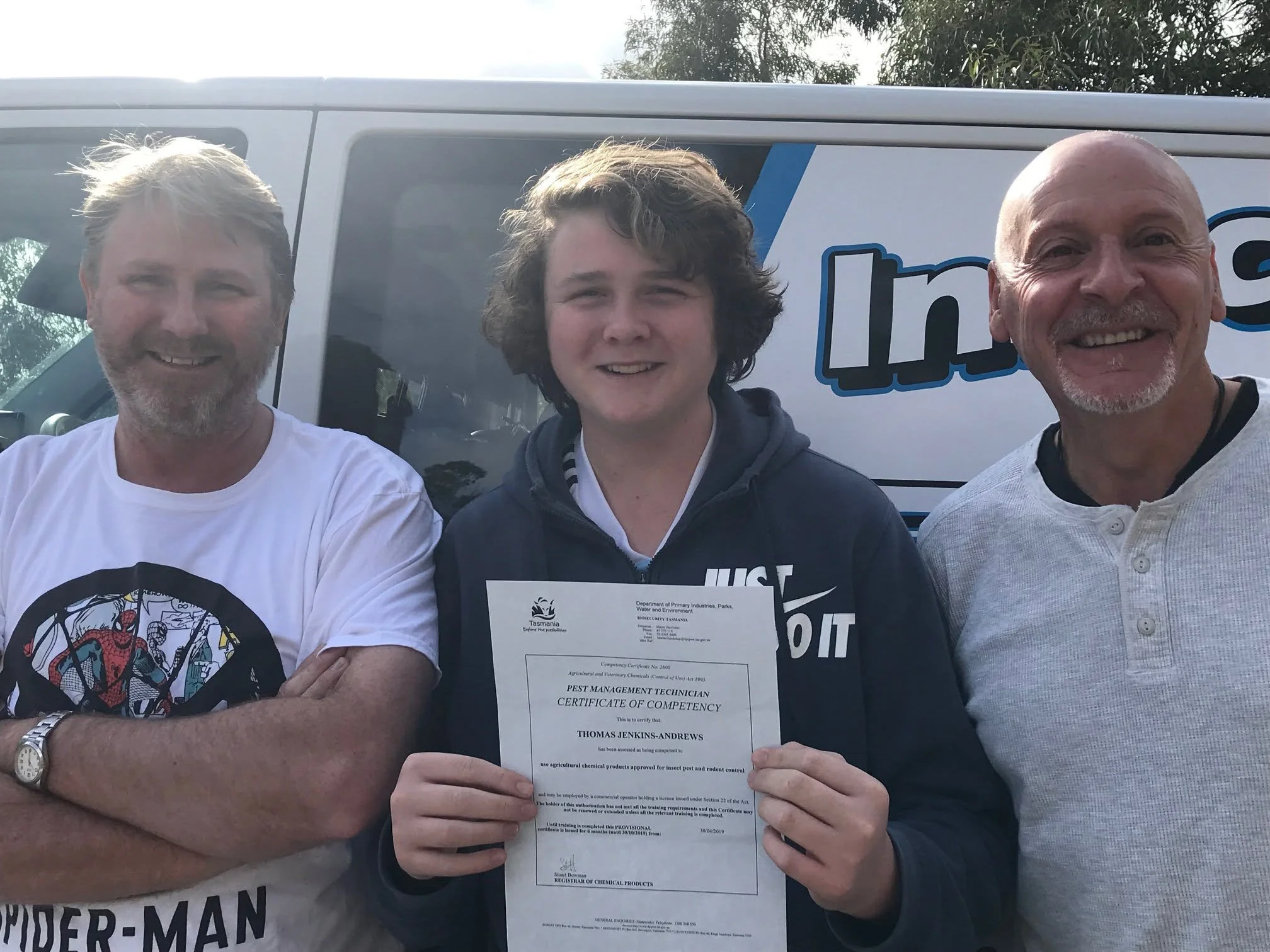 Three men standing outdoors in front of a vehicle with a partial logo. The man in the middle is holding a certificate of competency for a technical qualification, and the other two men are smiling.