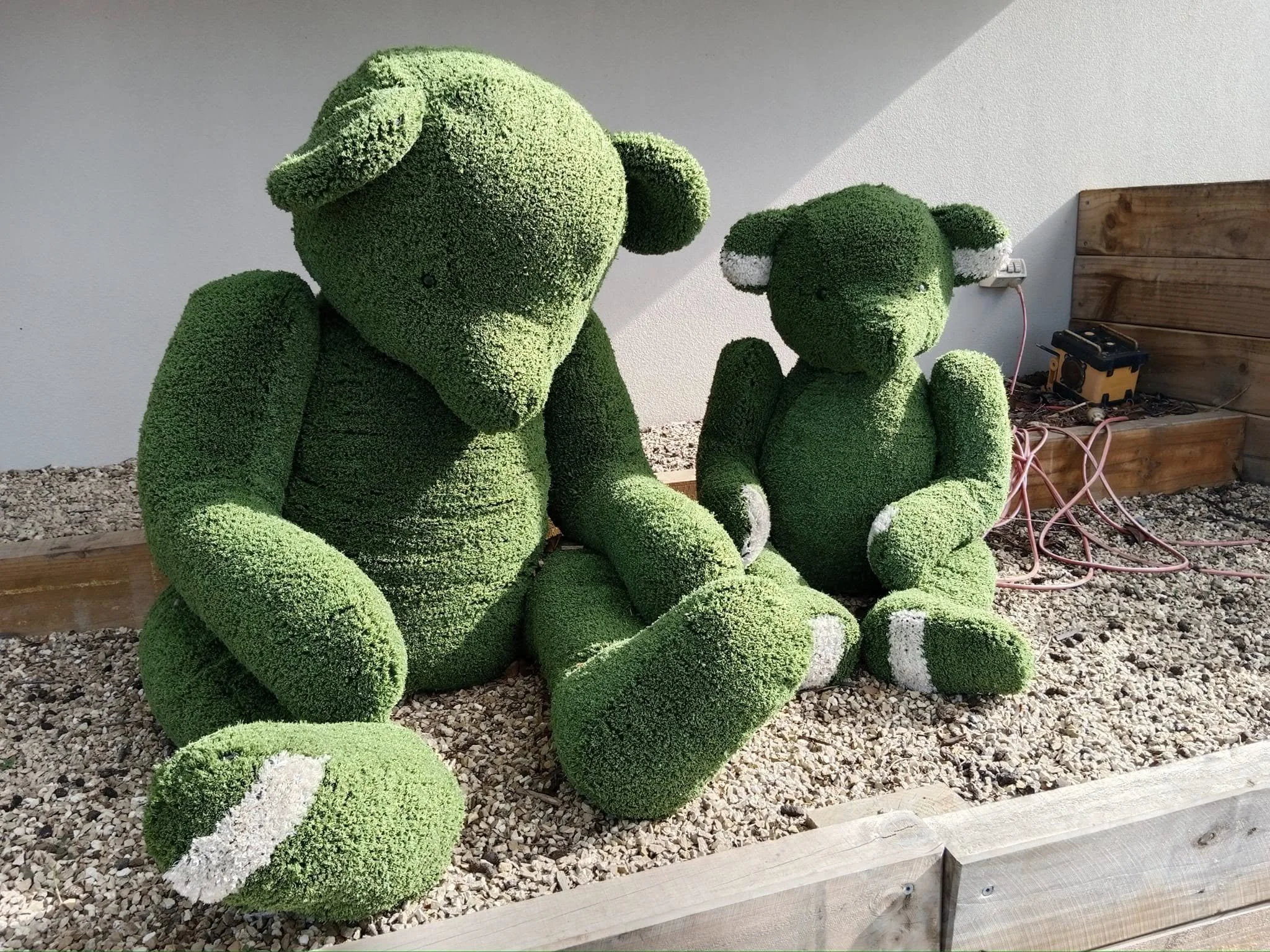 Two large topiary sculptures of dogs made from green plants, sitting on gravel beside a weathered wooden planter box with electrical equipment and pink cables on the ground.