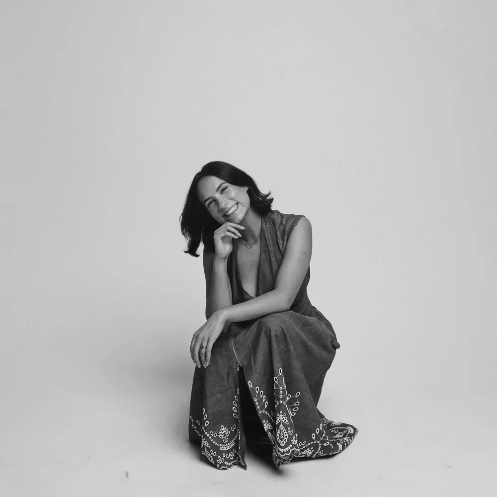 Black and white photo of a Rosemary Bashford with shoulder-length hair, sitting on the floor against a plain background, wearing a sleeveless top and wide pants with a decorative pattern.