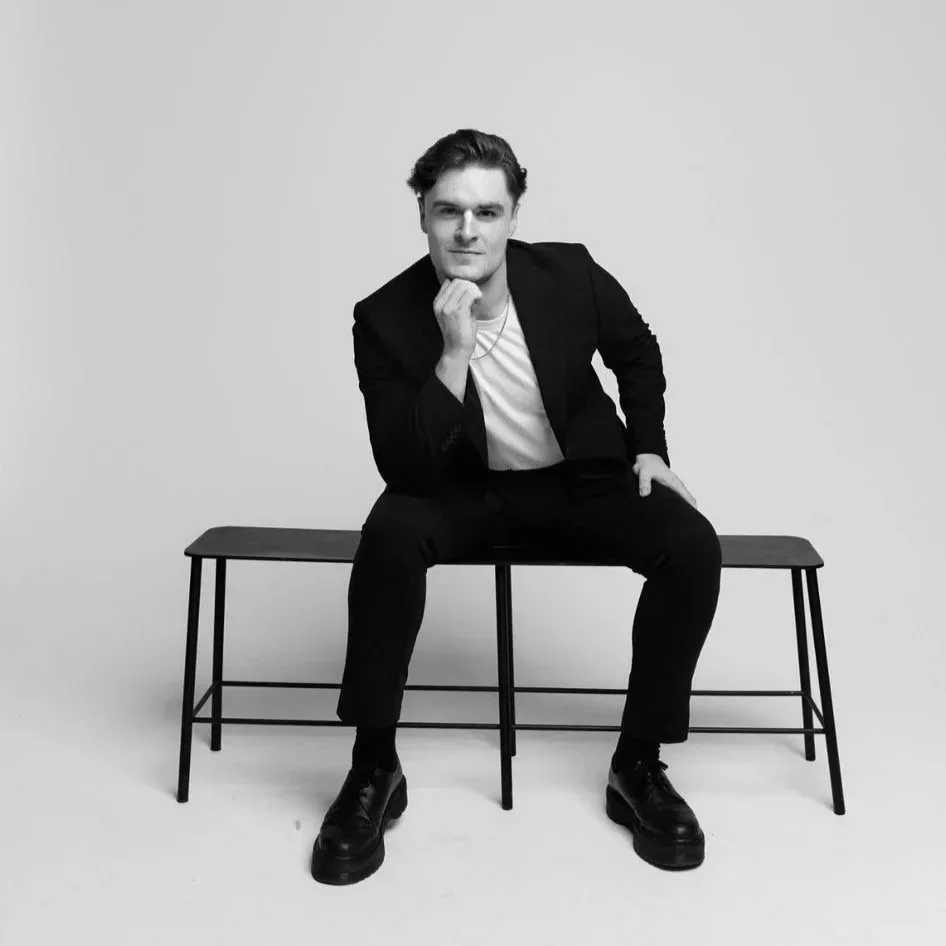 Flynn Starrs in a black blazer and white t-shirt sitting on a bench, resting his chin on his hand, against a plain background.