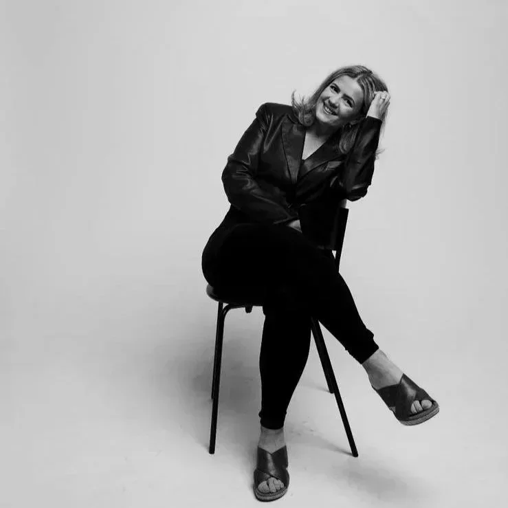 A woman sitting on a chair, smiling, wearing a dark leather jacket and black pants, with one hand touching her hair, in a black and white photo