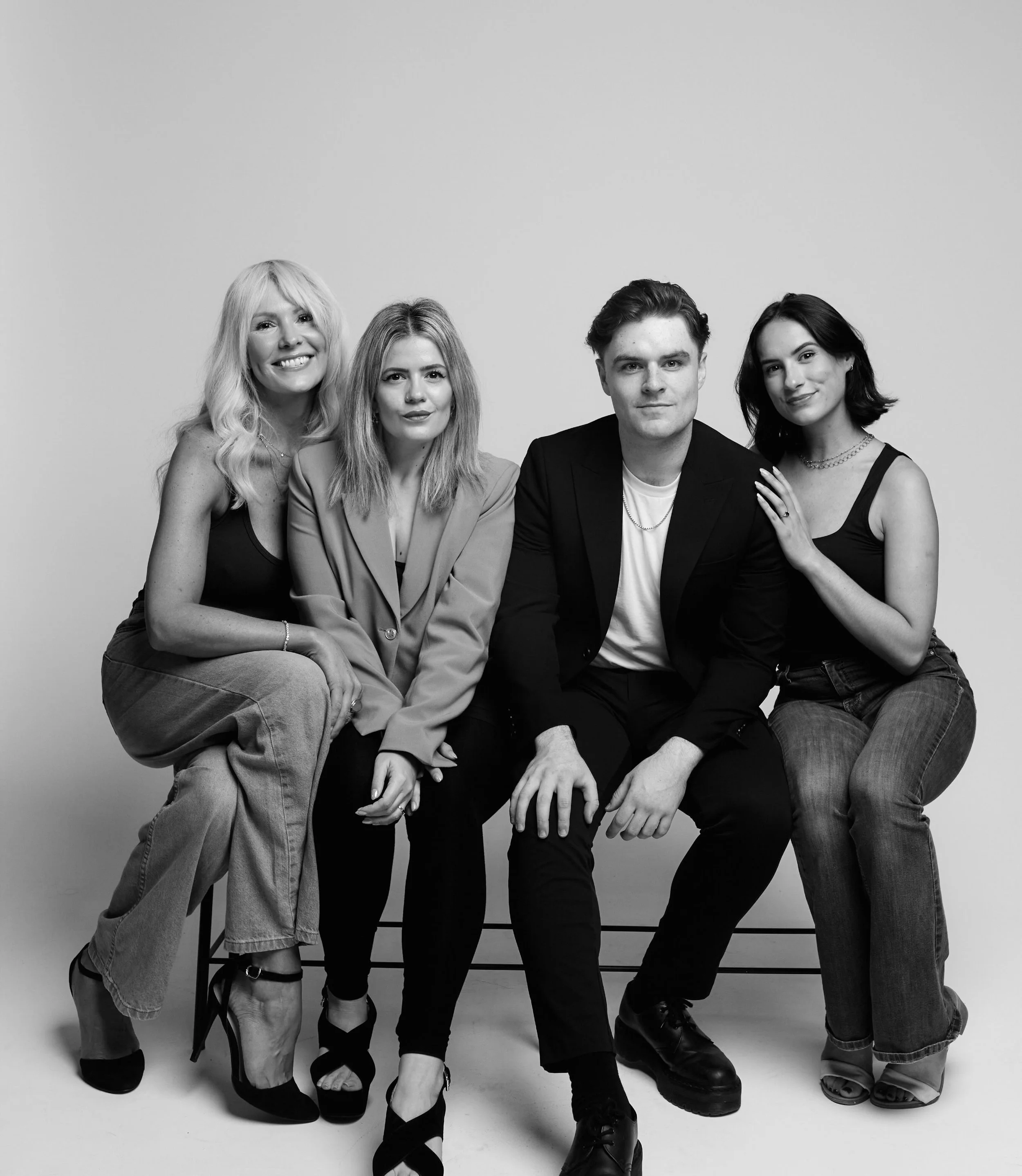 Black and white photo of four people sitting on chairs against a plain background. From left to right: a woman with long blonde wavy hair, wearing a sleeveless top and jeans, smiling; a woman with long straight light hair, wearing a blazer and dark pants, looking serious; a man with dark wavy hair, wearing a blazer and light shirt, looking at the camera; and a woman with shoulder-length dark hair, wearing a sleeveless top and jewelry, smiling.
