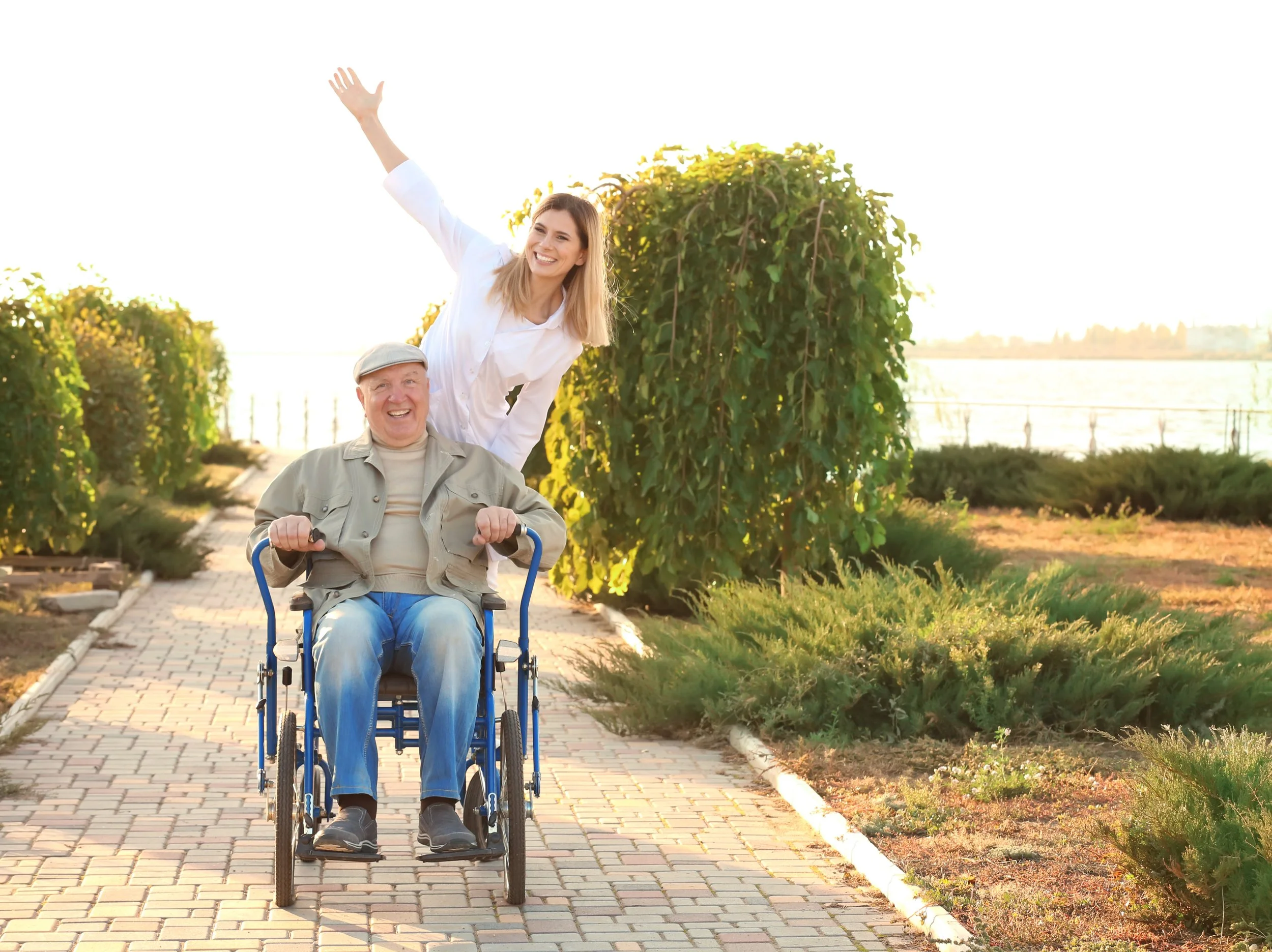 A young woman with a bright smile, being carried on the shoulders of an elderly man in a wheelchair along a paved path outdoors, with green bushes and a body of water in the background during sunset.
