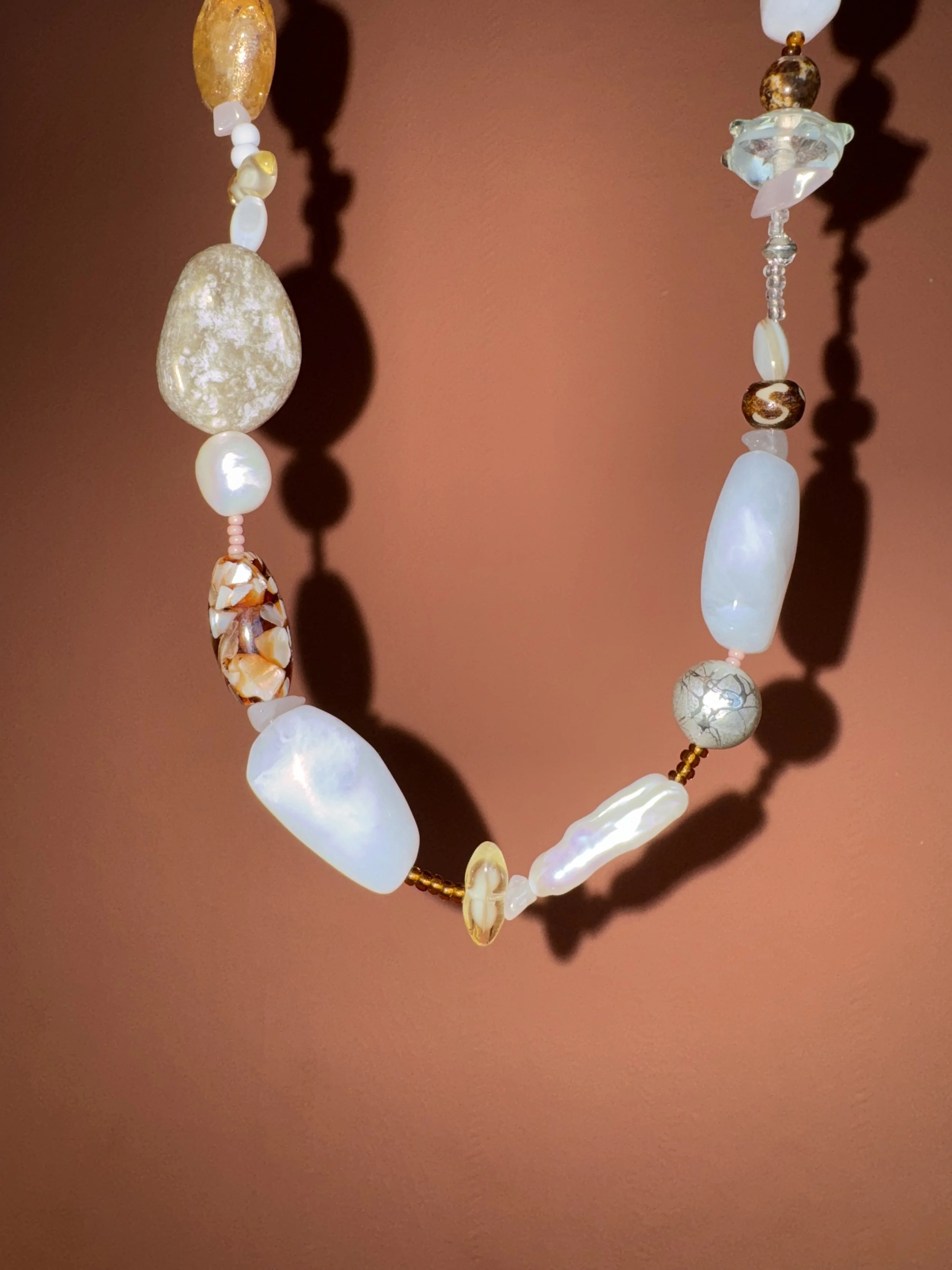 Close-up of a beaded necklace with white, pearl-like, and multicolored stones hanging against a reddish-brown background, casting shadows.