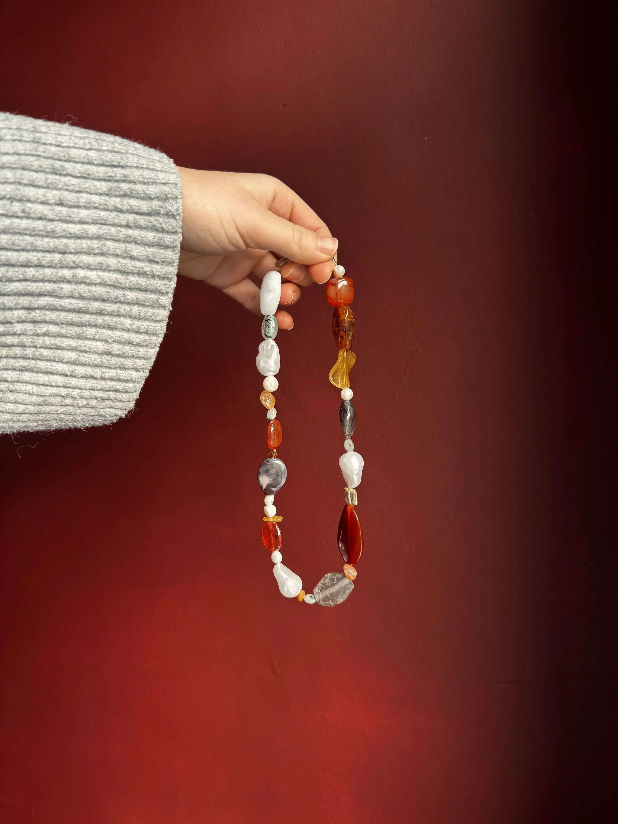 A hand holding a string of multicolored beads against a red background. The hand is partially covered by a gray, ribbed sweater.