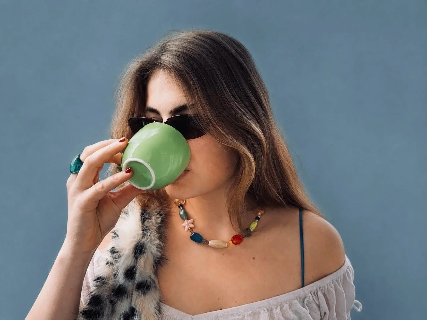 Woman with wavy brown hair wearing sunglasses and a colorful beaded necklace drinking from a green mug, standing against a plain blue background.