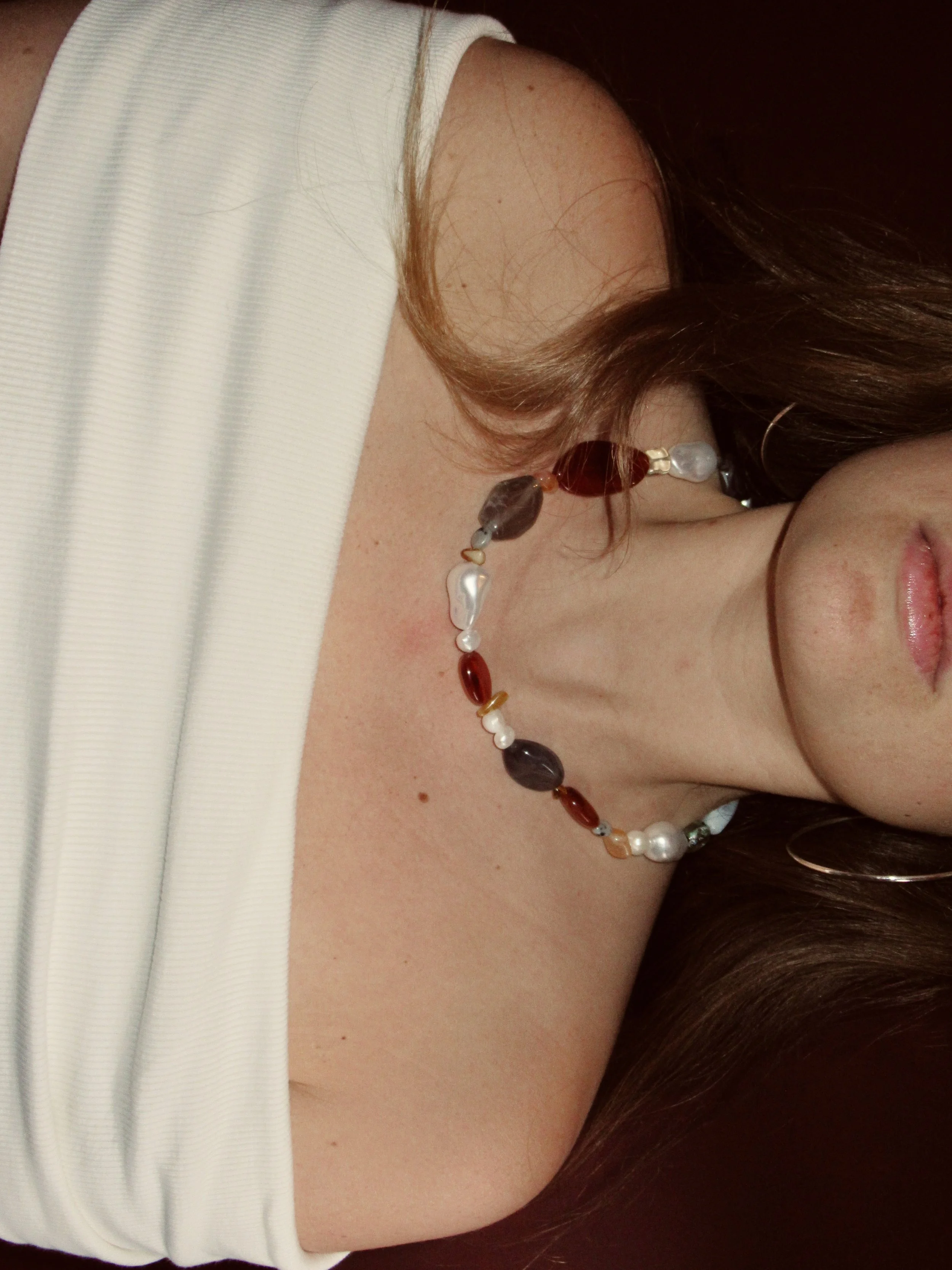 Close-up of a woman's shoulder and neck, wearing a white sleeveless top and a colorful beaded necklace, with long brown hair partially visible.