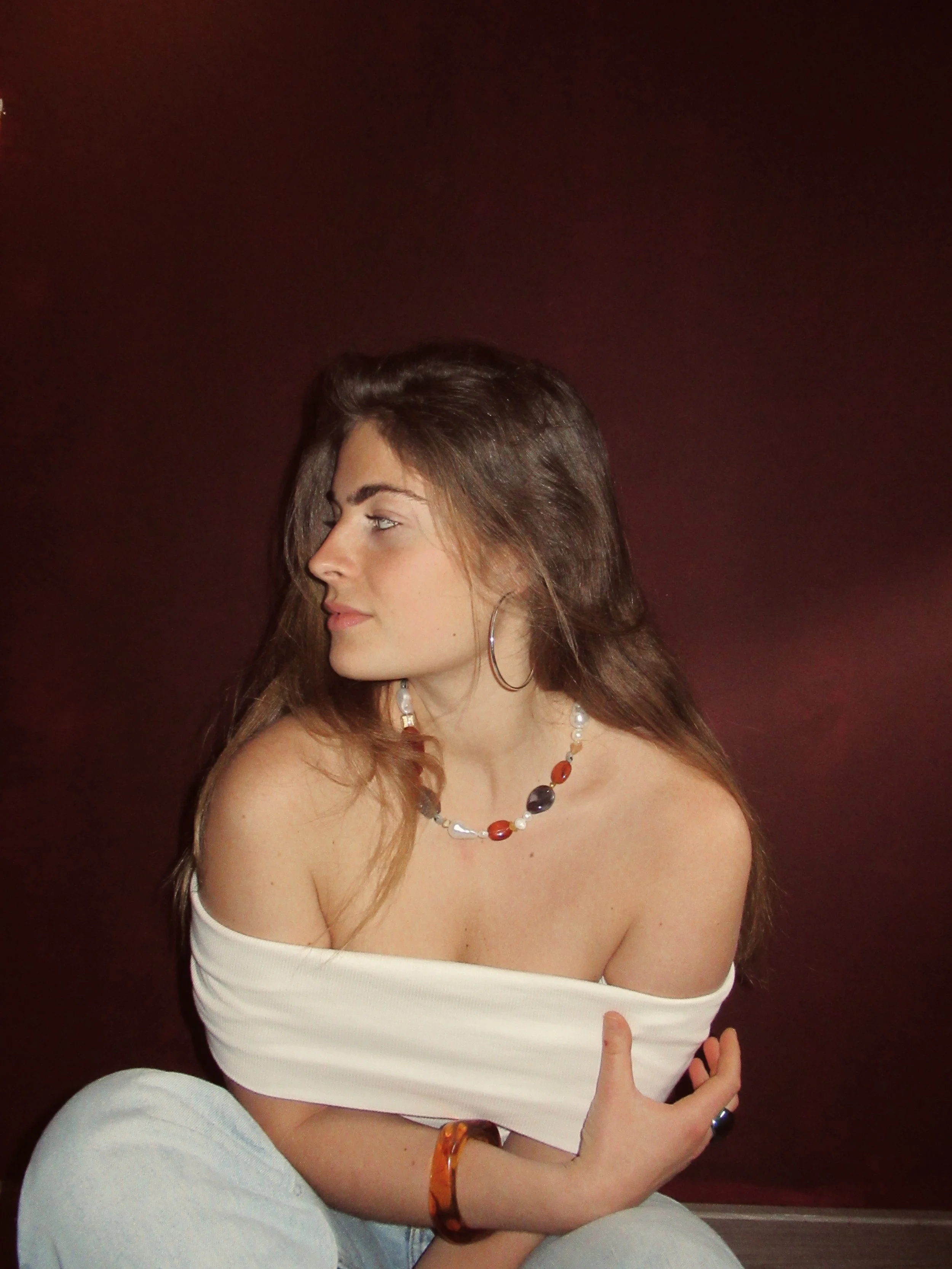 A young woman with long brown hair, wearing a white off-the-shoulder top, blue jeans, hoop earrings, and colorful jewelry, sitting against a dark red background, with her eyes closed and her right hand resting on her knee.