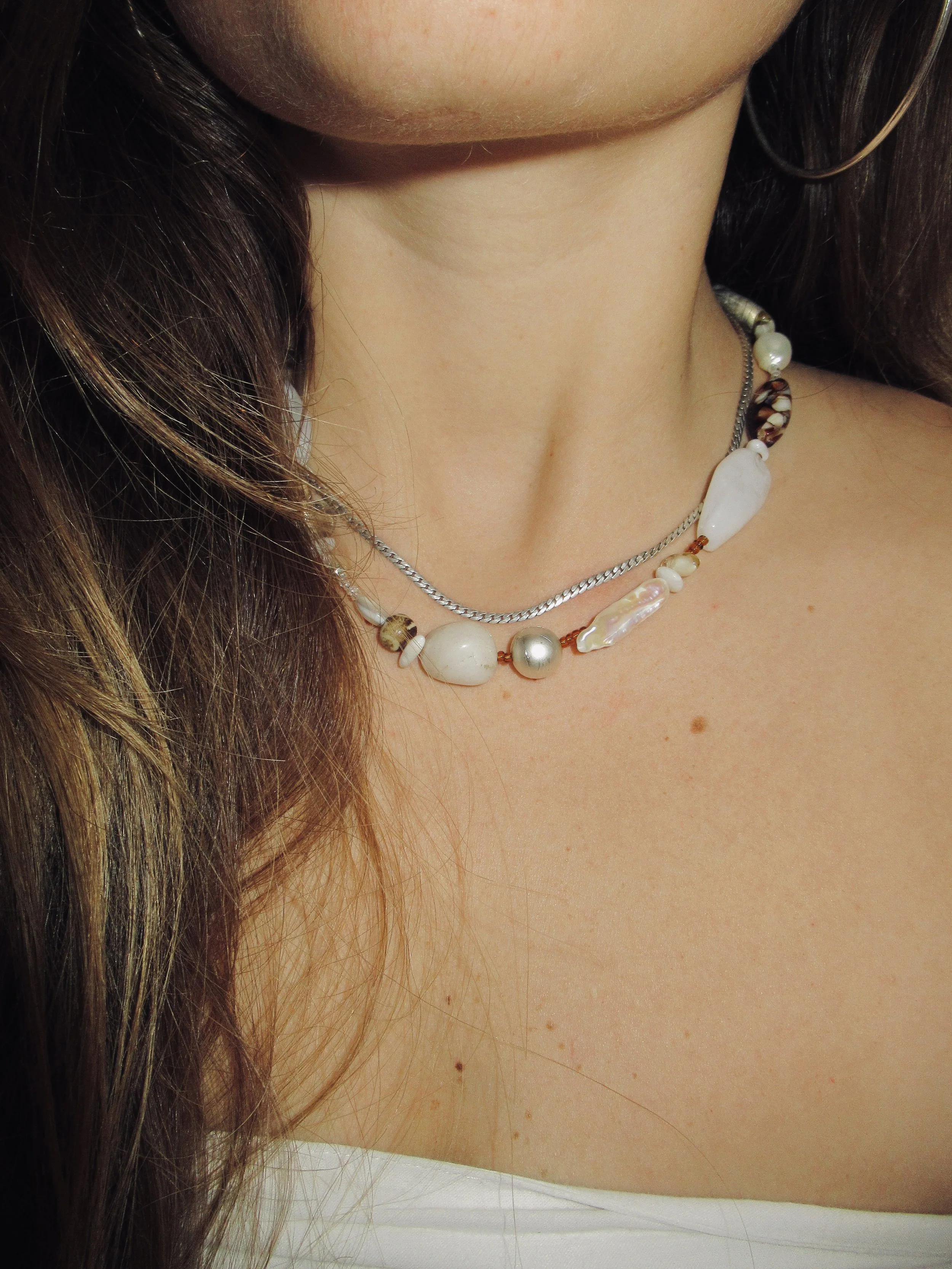 Close-up of a woman's neck and collarbone with layered necklaces, including pearls and shells. She has long, brown hair and wears hoop earrings.