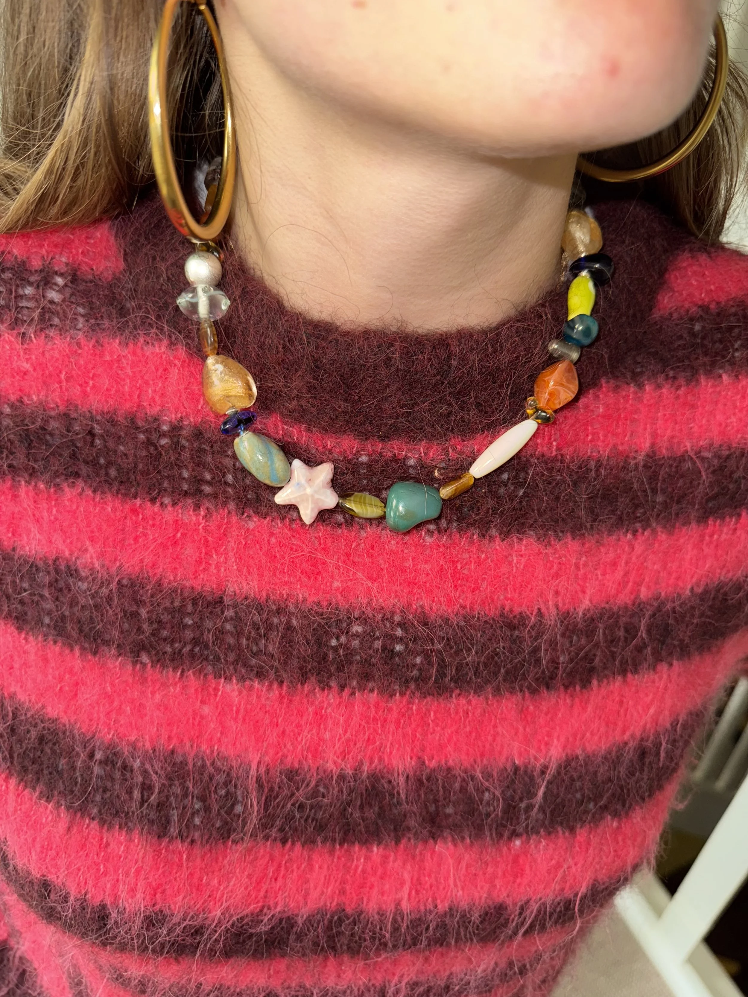 Close-up of a person wearing a red and black striped fuzzy sweater, large gold hoop earrings, and a colorful beaded necklace with a star-shaped pendant.