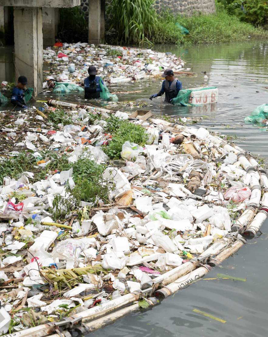 People cleaning up trash and plastic waste from a polluted water body, with a large amount of debris and garbage floating on the water and along the shoreline.
