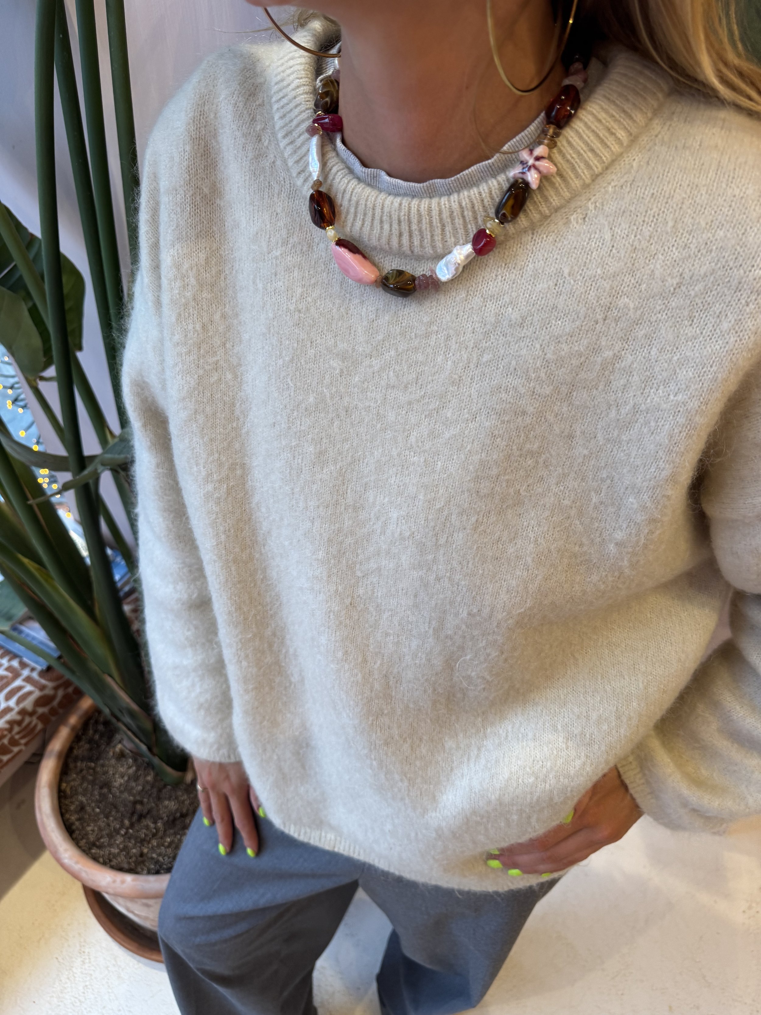 A woman wearing a beige sweater, gray pants, and neon green nail polish, standing indoors near a potted plant.