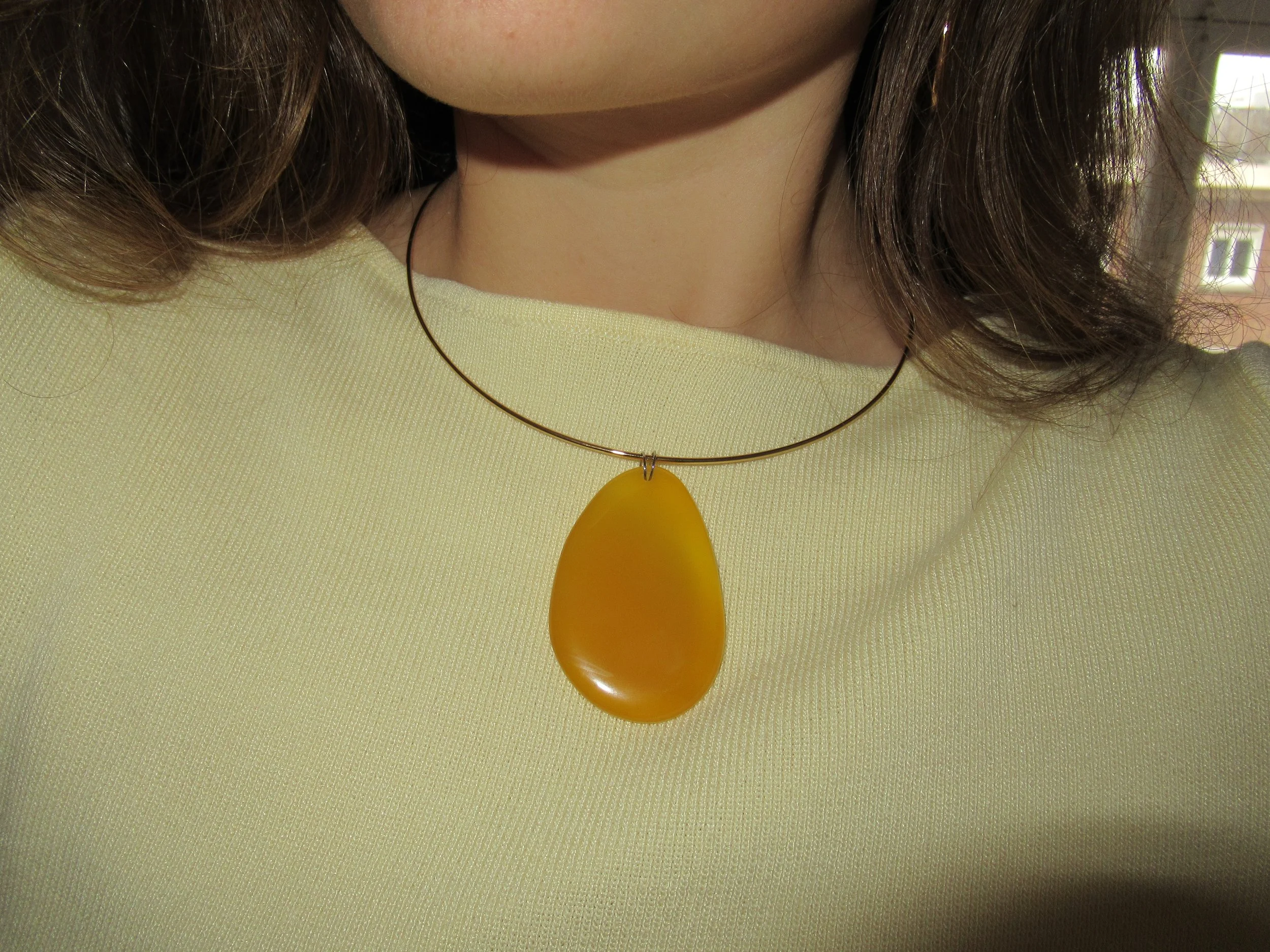 Close-up of a woman's neck wearing a yellow top, a gold necklace with a yellow pendant, and brown, wavy hair with a window in the background.