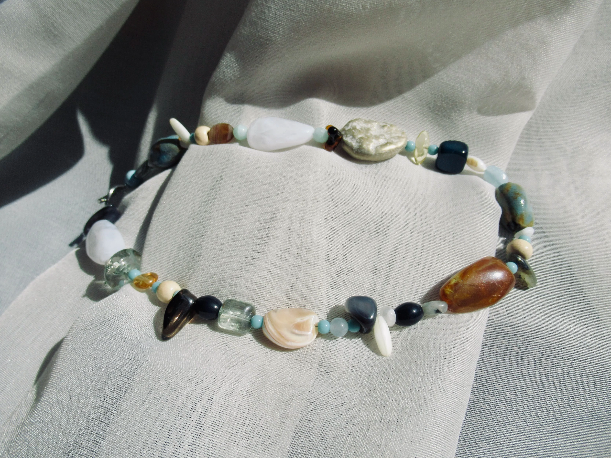 A beaded necklace with various stones and shells laid out on a light-colored fabric surface.