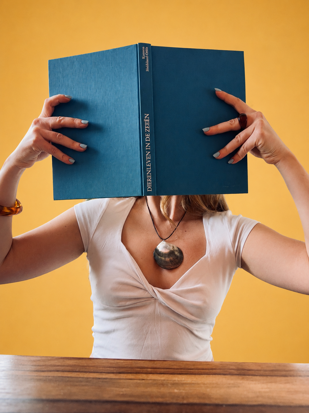 Person holding a blue hardcover book in front of their face, with a yellow background and a wooden table in the foreground. The person wears a white top, a large shell necklace, and has painted nails with a ring on the finger.