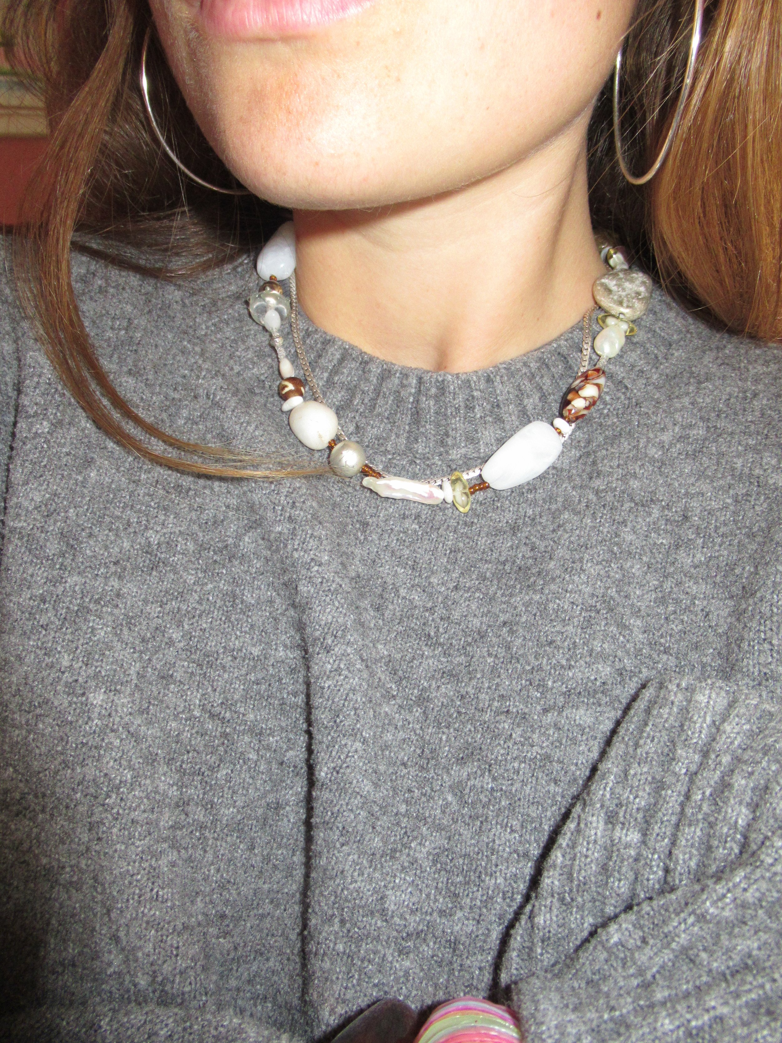 Close-up of a woman wearing a gray top, pearl and shell necklace, silver hoop earrings, and a pink ring.