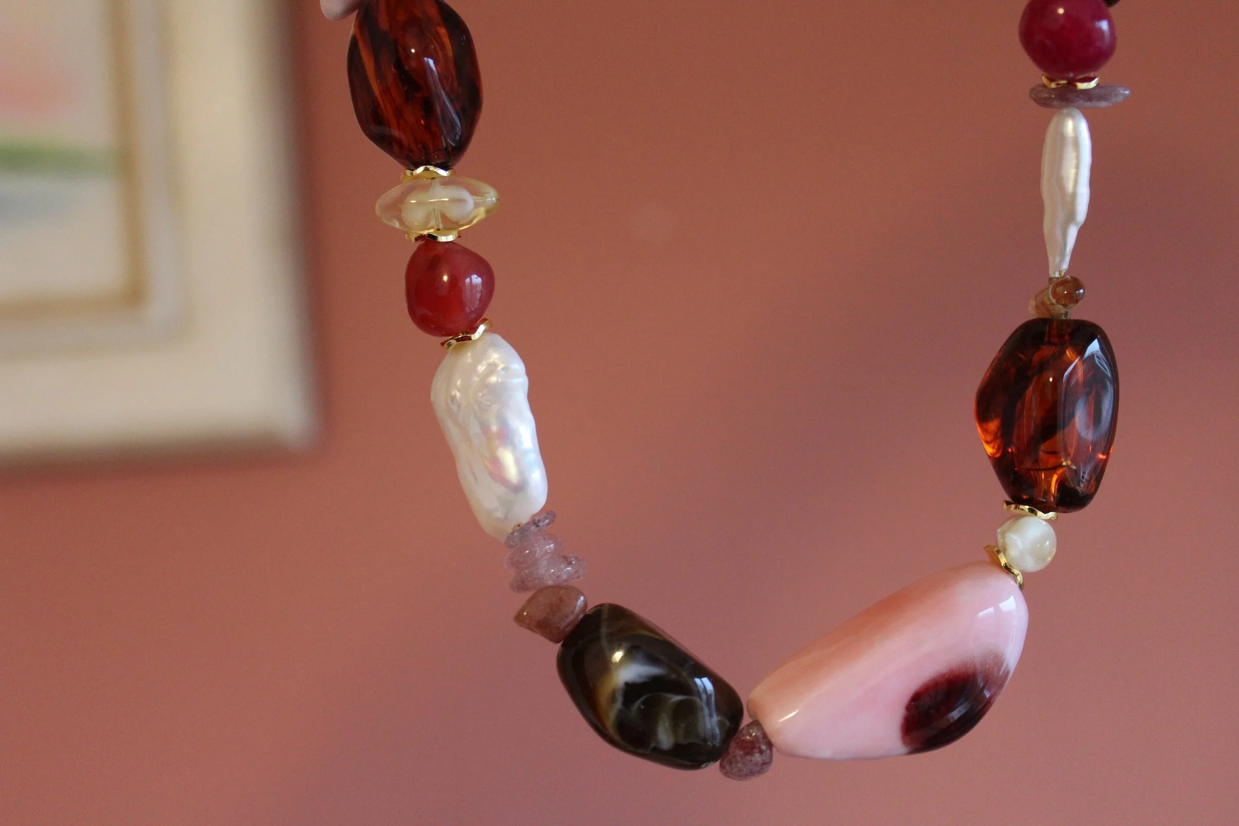 Close-up of a string of various beads, including pearls, red and black stones, and other semi-precious stones, held by a person's hand against a blurred background.