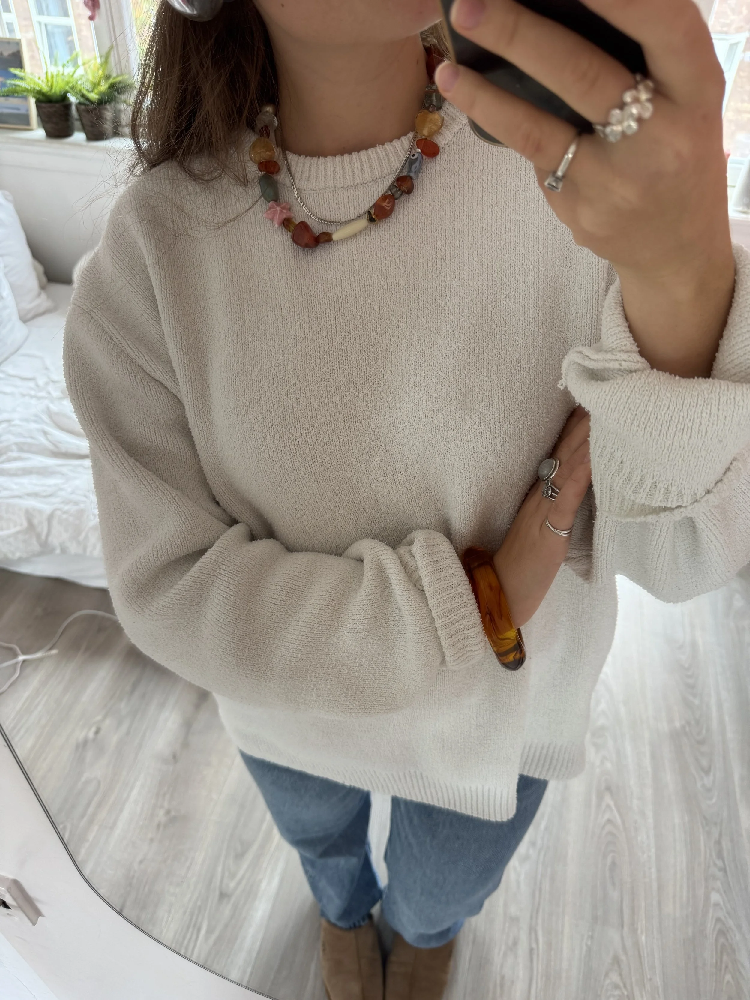 Person taking a mirror selfie wearing a cream-colored sweater, blue jeans, and jewelry including necklaces and rings, in a room with a wooden floor, white walls, and potted plants on the window sill.