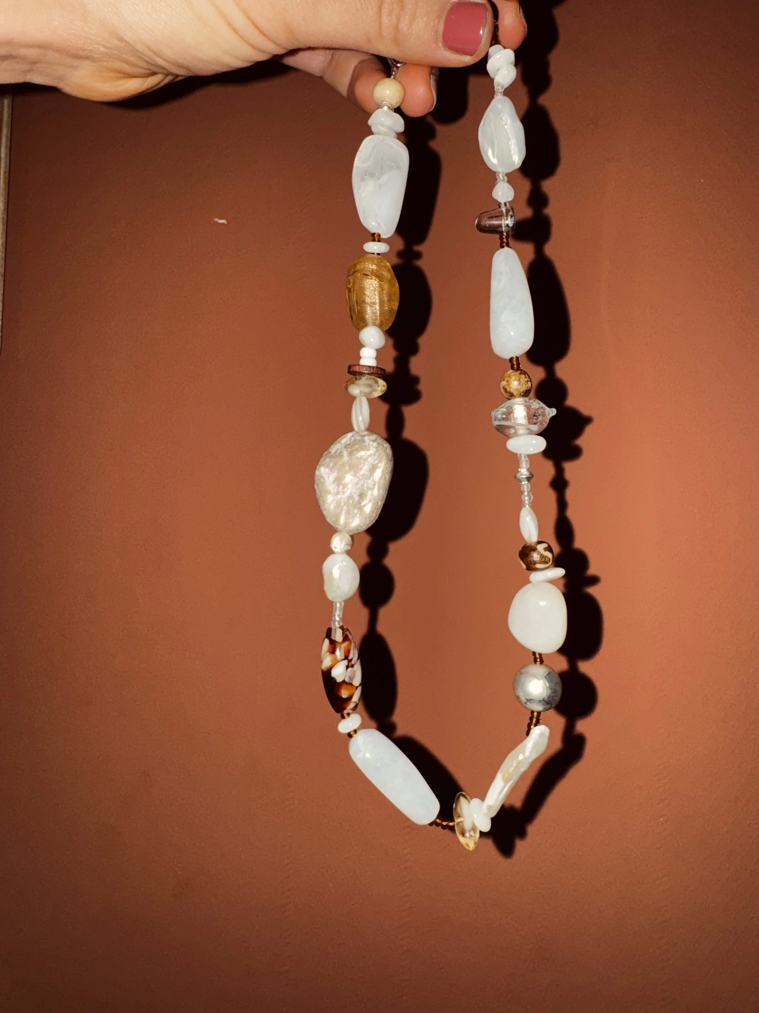A hand holding a beaded necklace with various white, brown, and translucent stones against a brown background.