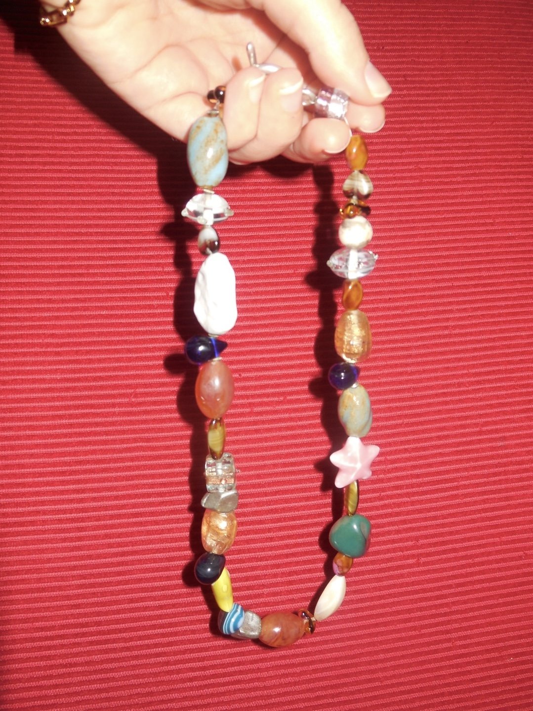 Colorful beaded necklace with various shapes and sizes of beads, held in a person's hand against a red textured fabric background.
