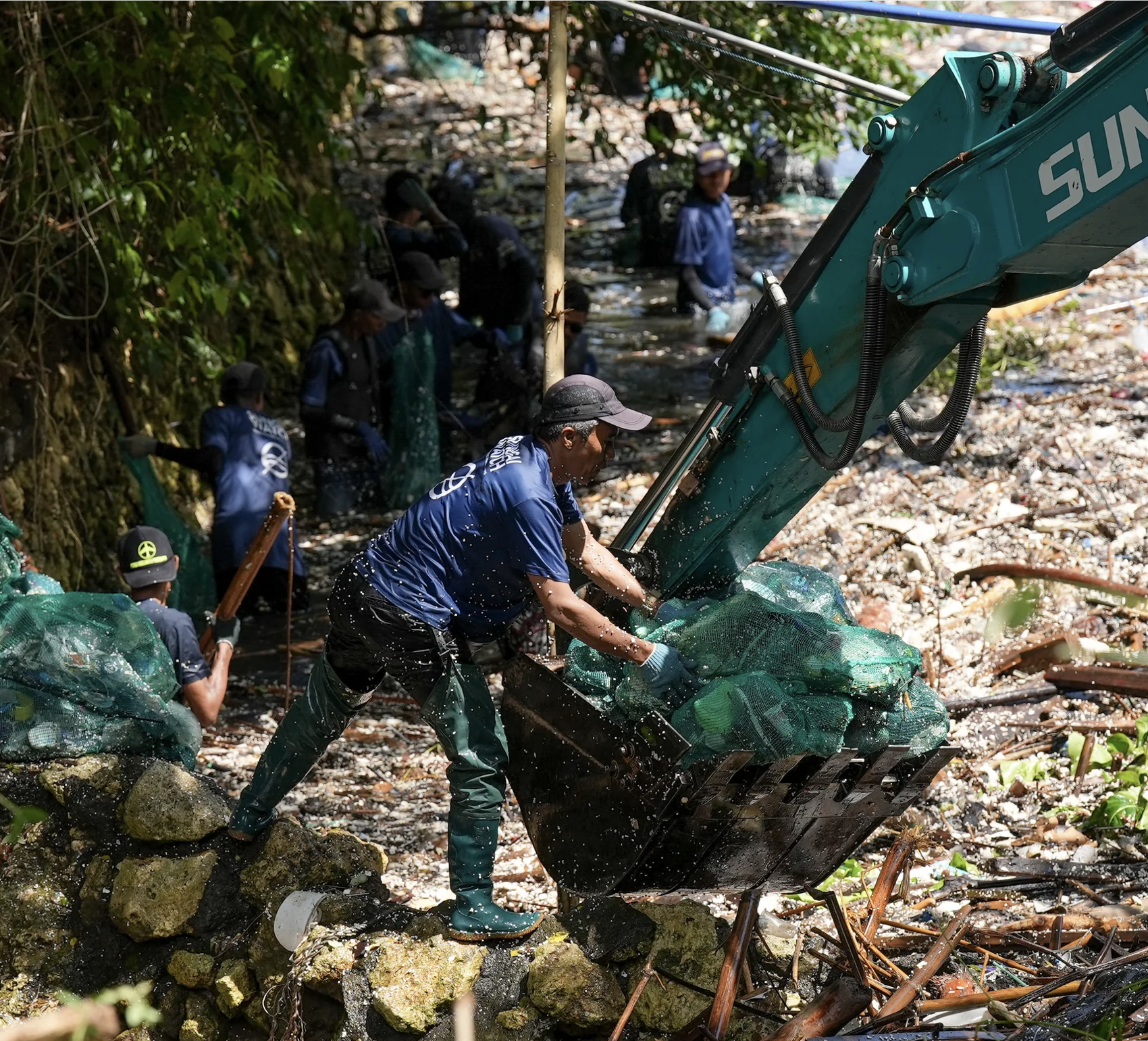 People working with a backhoe to clear debris and trash from a river, surrounded by rocks, green vegetation, and fallen leaves.