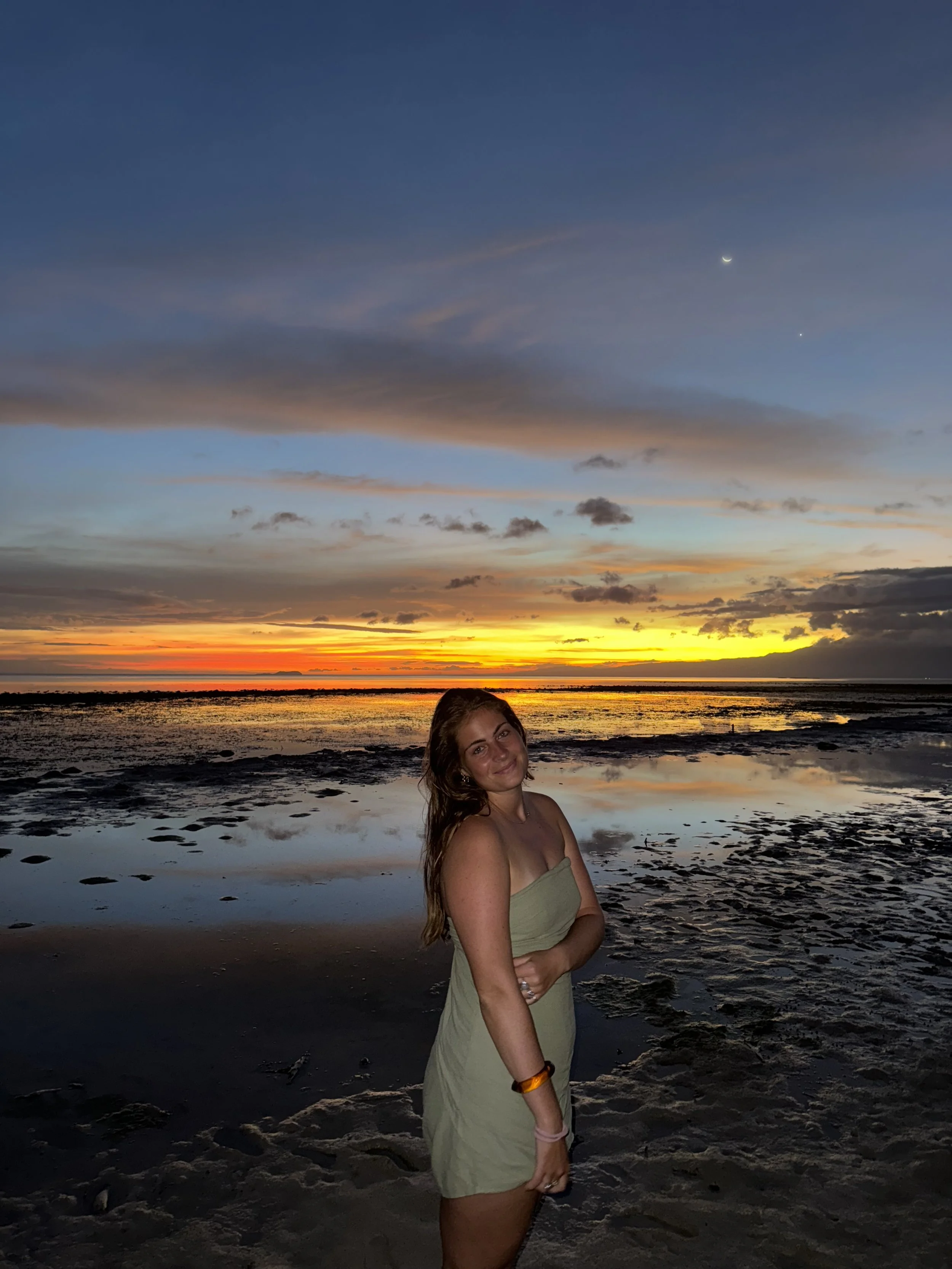 A woman in a strapless dress standing on the beach during sunset with a colorful sky, clouds, and the moon visible in the background.