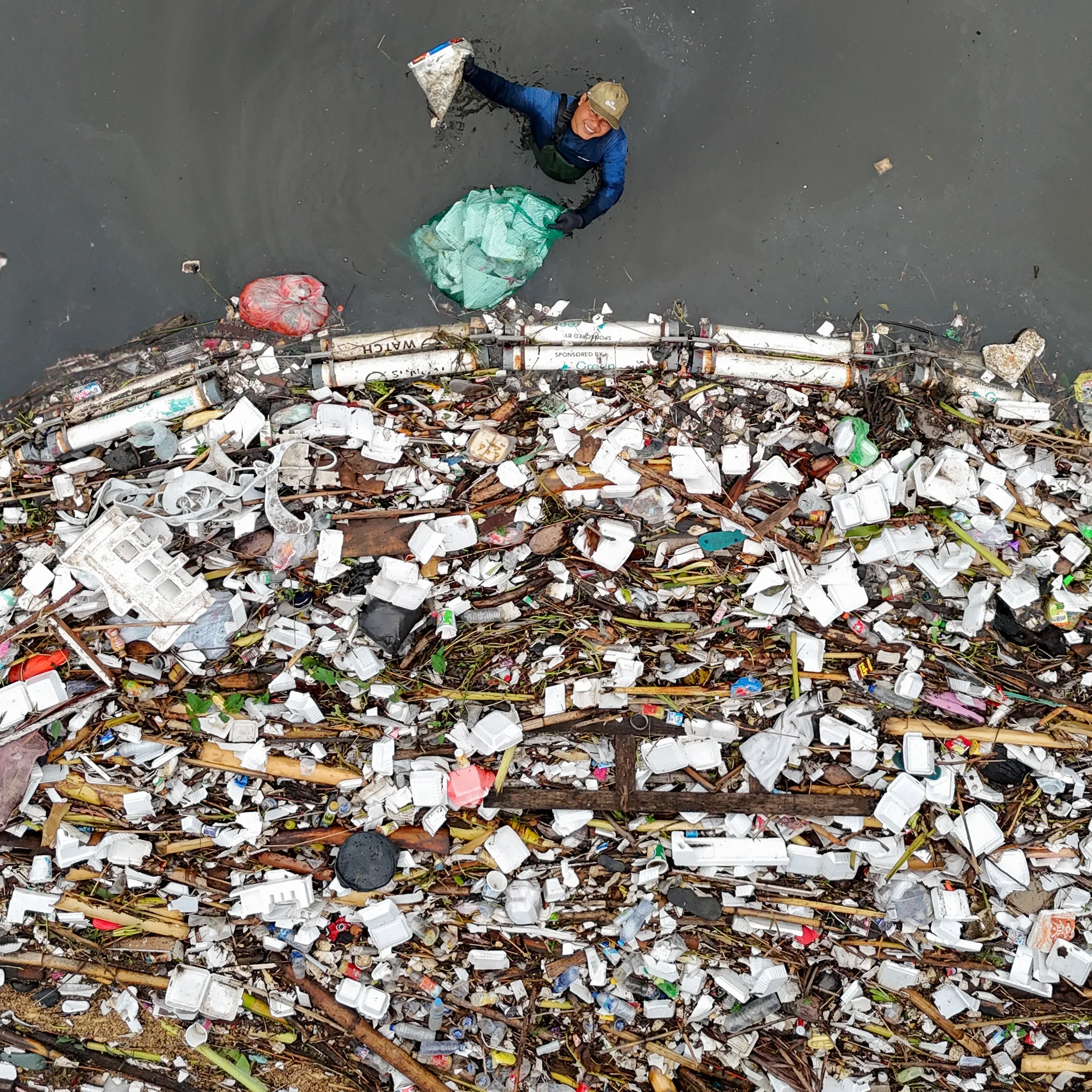 A person in a blue jacket and a tan cap is standing in a muddy water body, picking up trash with a black glove. The water is filled with considerable trash, including plastic debris, foam, and trash floating on the surface and along the muddy shoreline.