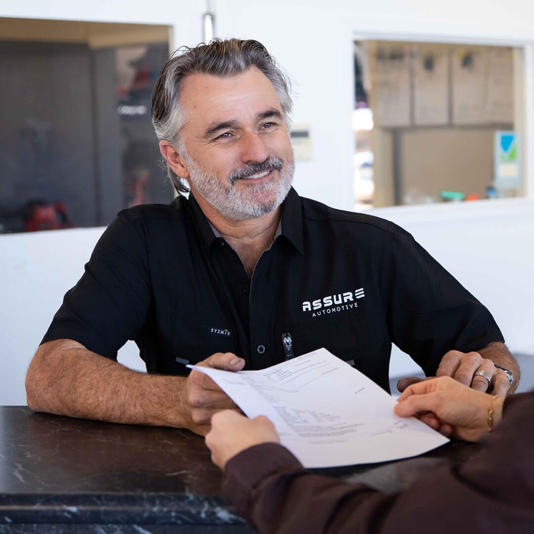 A smiling man with gray hair and beard wearing a black shirt with 'Assure Automotive' logo, passing a paper to someone across a table in a mechanic service centre.