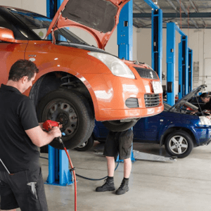 Mechanic working on an orange car lifted on a hydraulic lift in an auto repair shop, with another vehicle in the background.