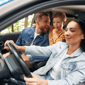 A woman driving a car with a man and a child smiling behind her, outdoors with trees visible through the car window.