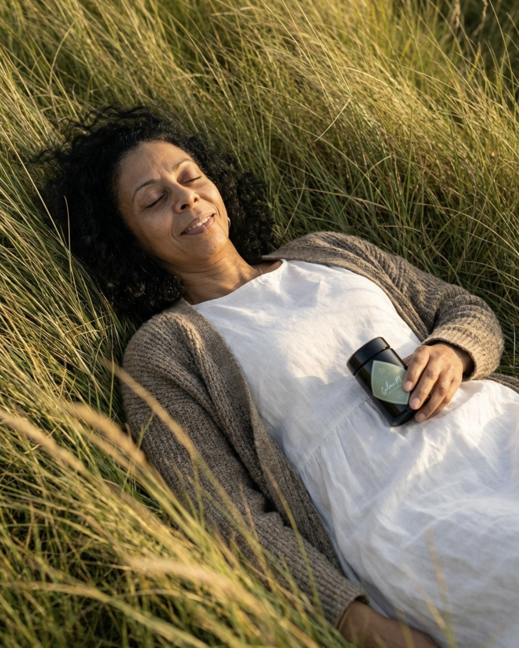 A woman relaxing on a grassy field, lying on her back with eyes closed and holding a travel mug.