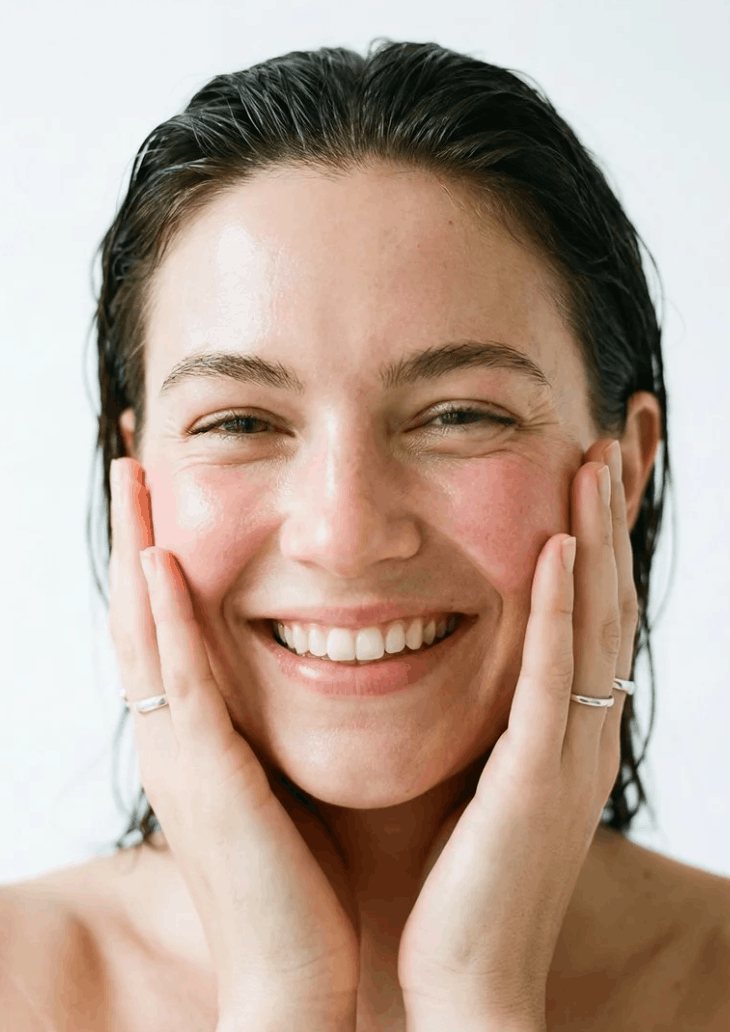 A smiling woman with wet, dark hair holding her face with both hands.