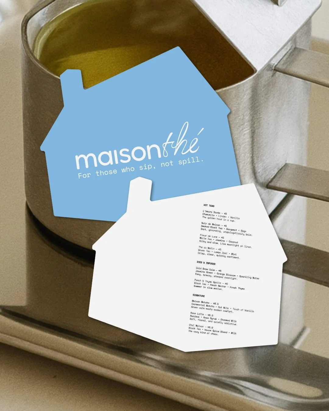 A menu for Maison Thé, a tea shop, placed on top of a tea sampler box, both resting on a silver surface near a set of stairs.