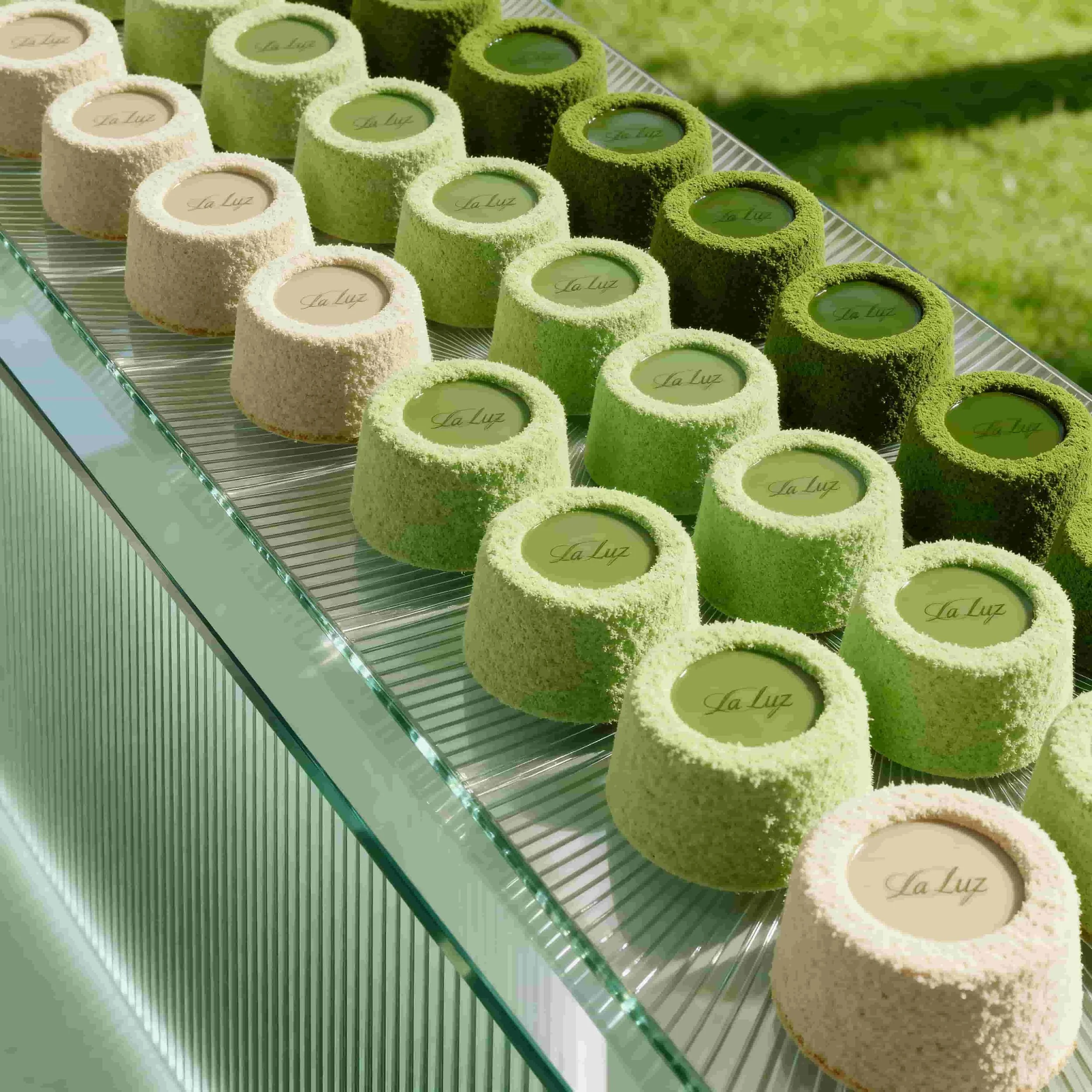 Rows of round desserts with green and white coconut coatings, each with a transparent top displaying the words 'La Luz', arranged on a glass tray outdoors.