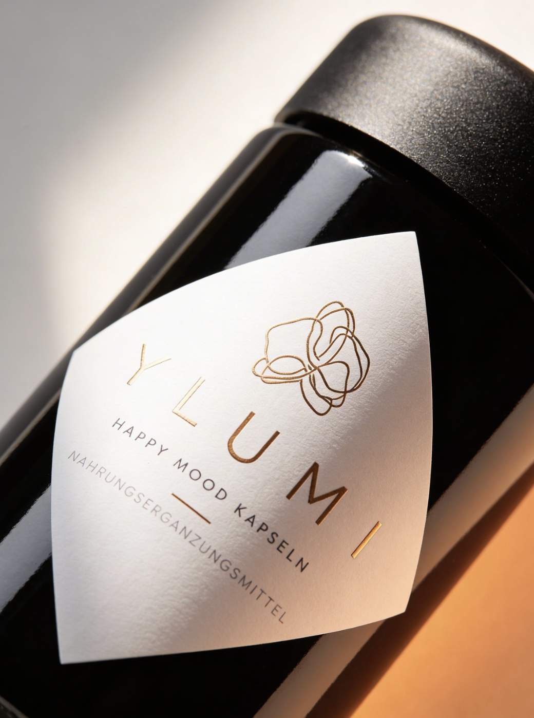 Close-up of a black bottle with a white label featuring a minimalist line drawing of a rose, and the text "Y L U M I" in gold lettering, with additional German text below.